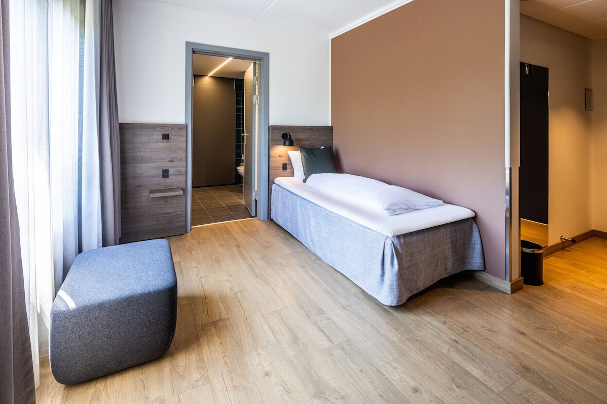 A modern hotel room with a single bed, a small seating area, and a bathroom visible through an open door.