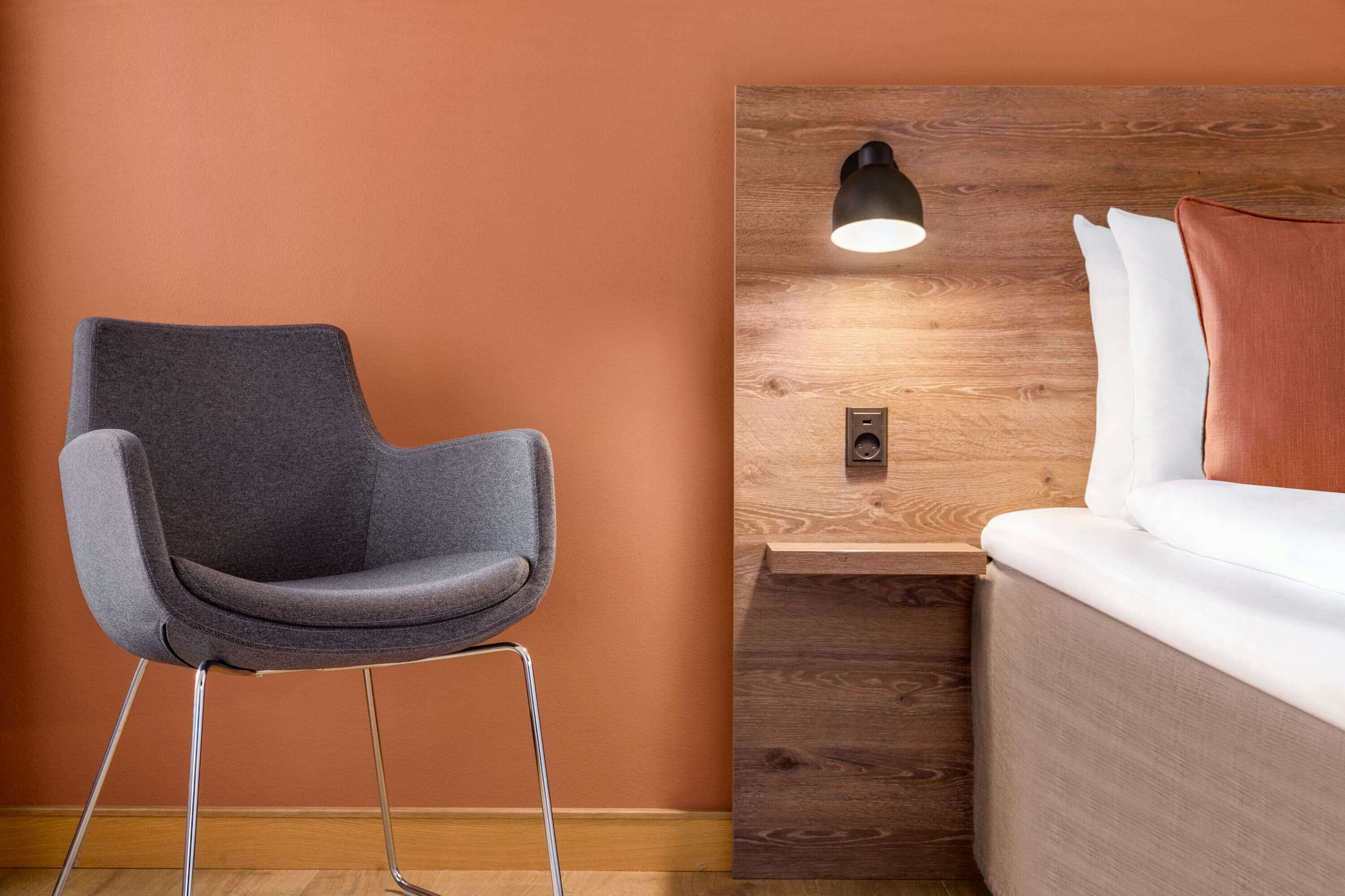 Une chambre moderne avec un fauteuil gris, une tête de lit en bois et une table de chevet avec une lampe.