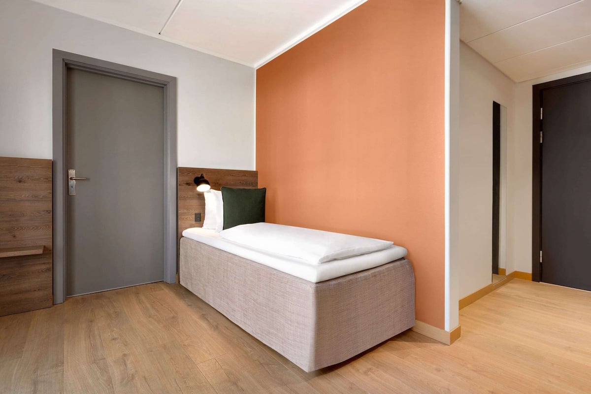 A small, modern room with a bed, a door, and a wall with a wooden panel and a green pillow.