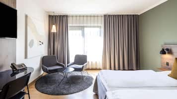 Une chambre d’hôtel comprenant un lit, deux chaises, une petite table, un bureau avec un téléphone, un téléviseur fixé au mur et une fenêtre avec des rideaux.