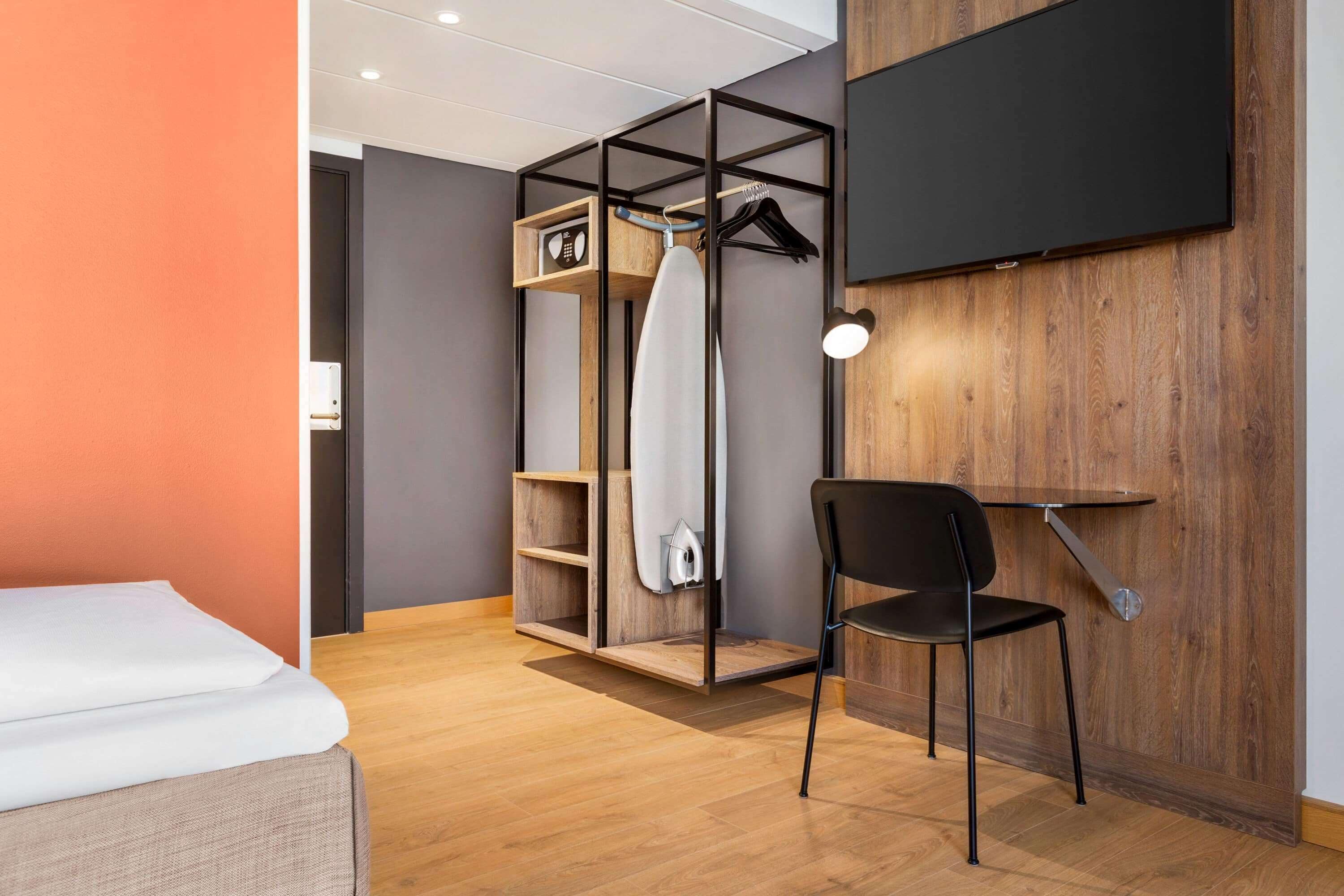 A modern hotel room with a bed, a wooden wardrobe, a chair, and a TV mounted on the wall.