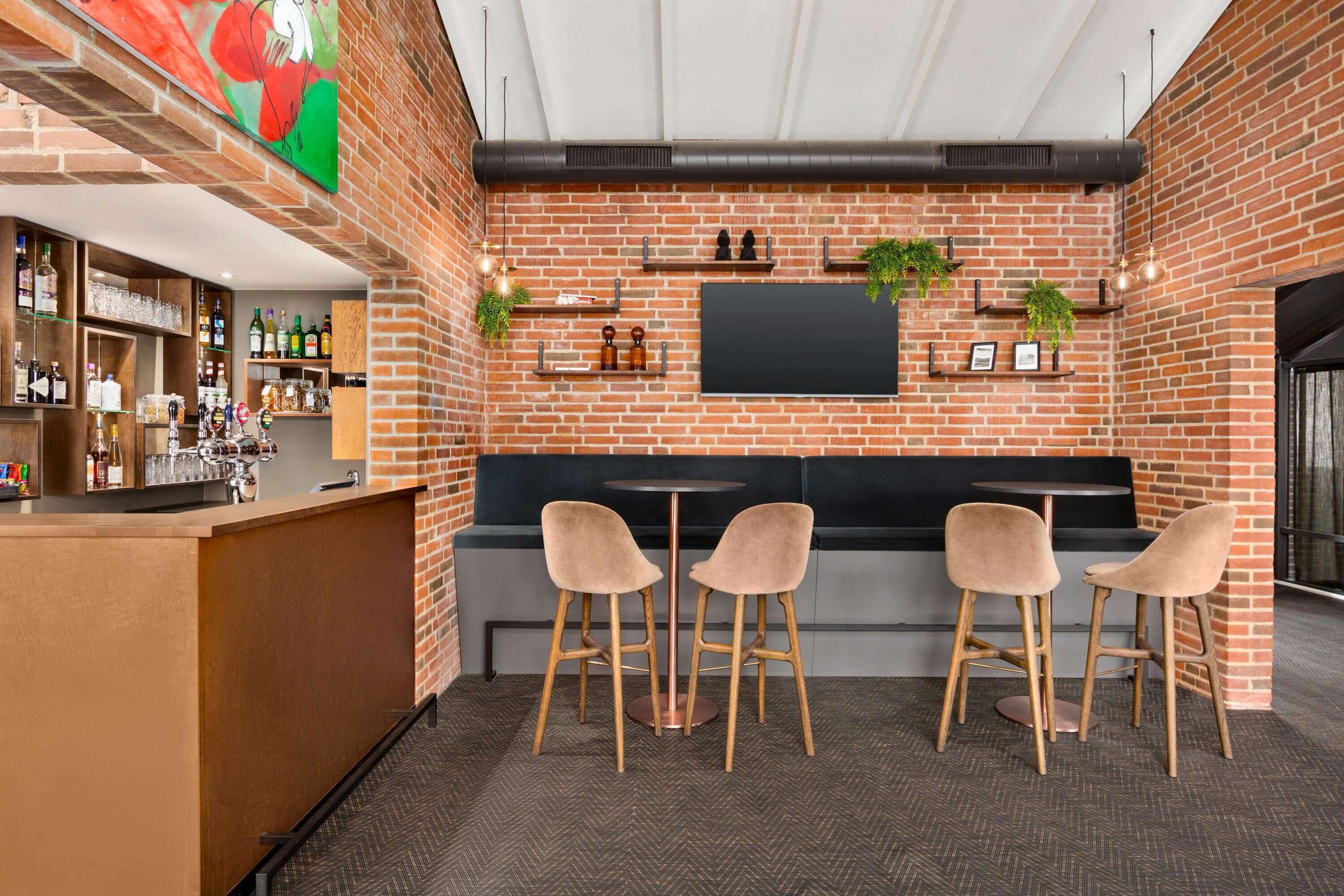 A bar area with a brick wall, shelves with bottles, a flat-screen TV, and four bar stools.
