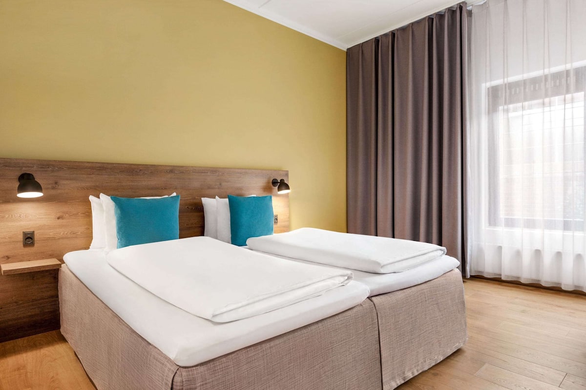 A double bed with white linens, two blue pillows, a wooden headboard, and a bedside table with a lamp.