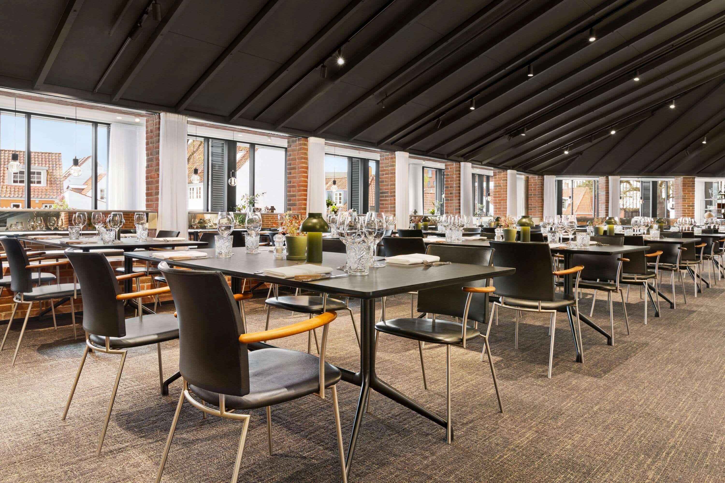 A modern dining area with multiple tables and chairs, large windows, and a brick wall.
