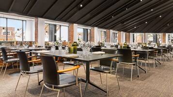A modern dining area with multiple tables and chairs, large windows, and a brick wall.