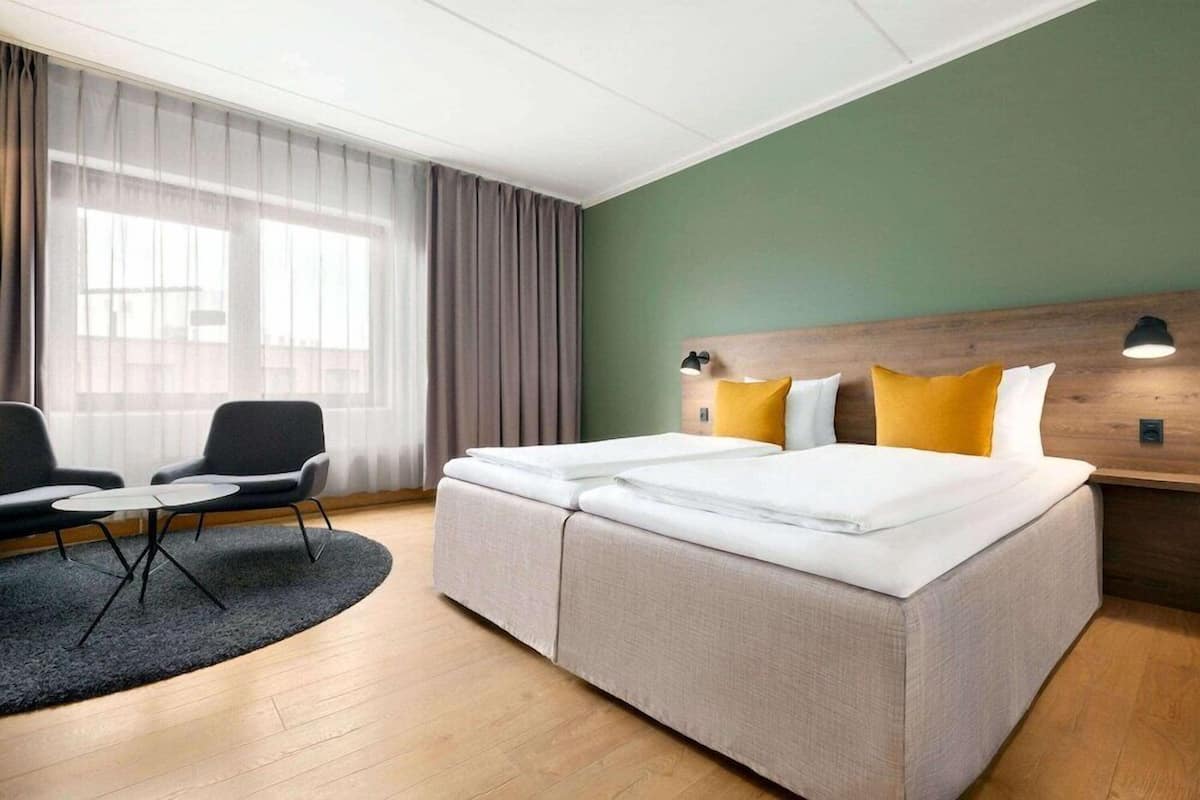 A hotel room with a large bed, a small table, two chairs, a wooden headboard, and a large window with curtains.