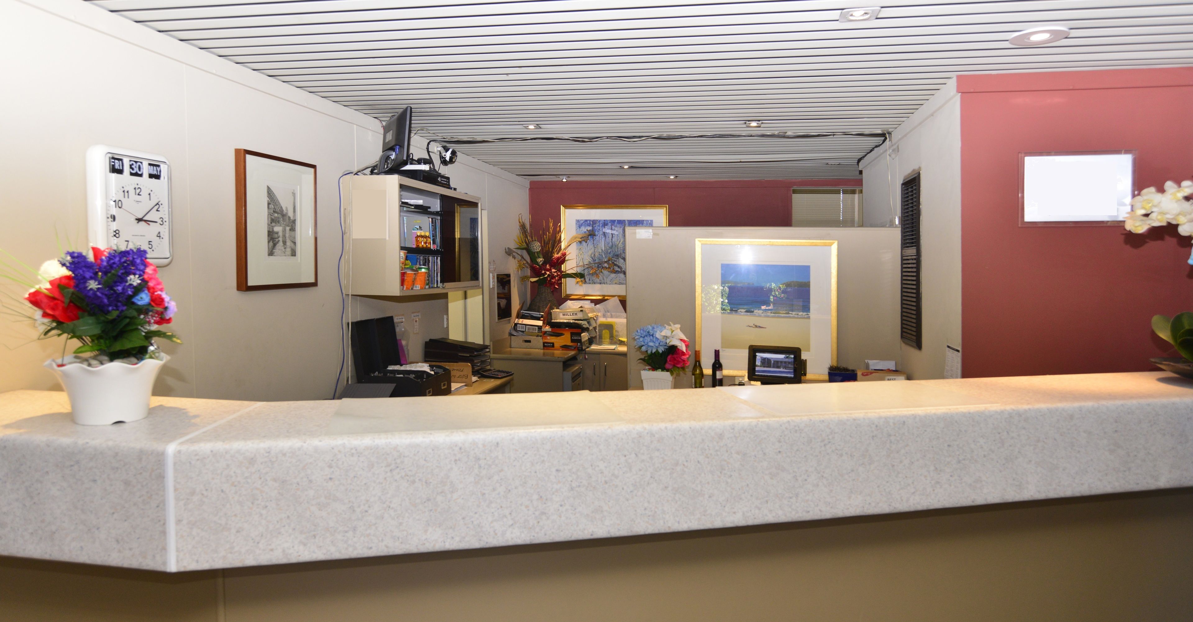 A reception area with a counter, a clock, framed pictures, and a small desk with a computer and a vase of flowers.
