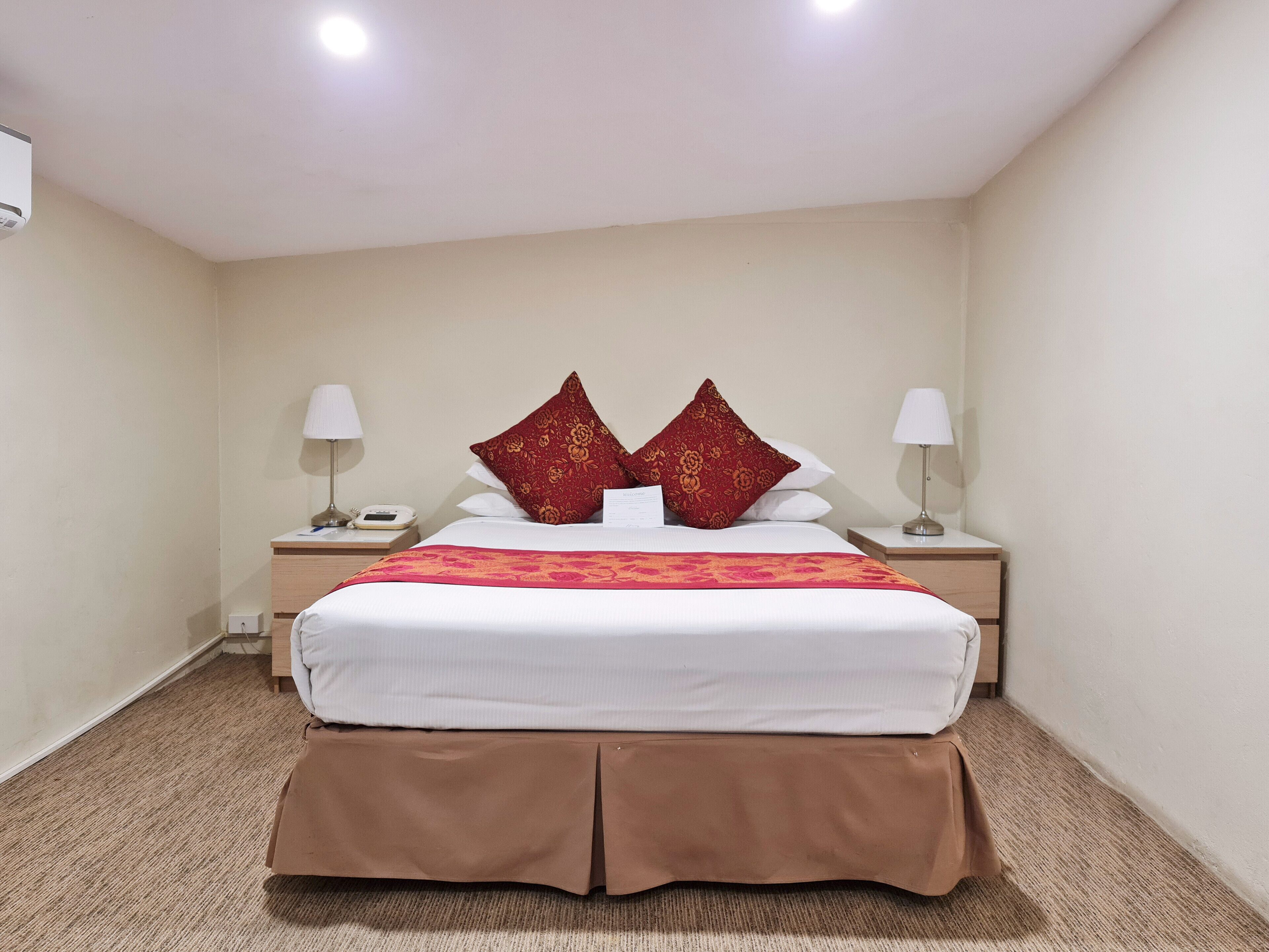 Une chambre d’hôtel avec un grand lit, deux tables de chevet, un couvre-lit à motifs rouges et blancs, et une base de lit marron avec une jupe.