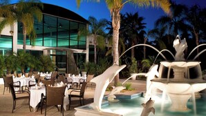 An outdoor dining area with tables and chairs, a fountain with dolphin sculptures, and a modern building in the background. - Four Points by Sheraton San Diego (San Diego)