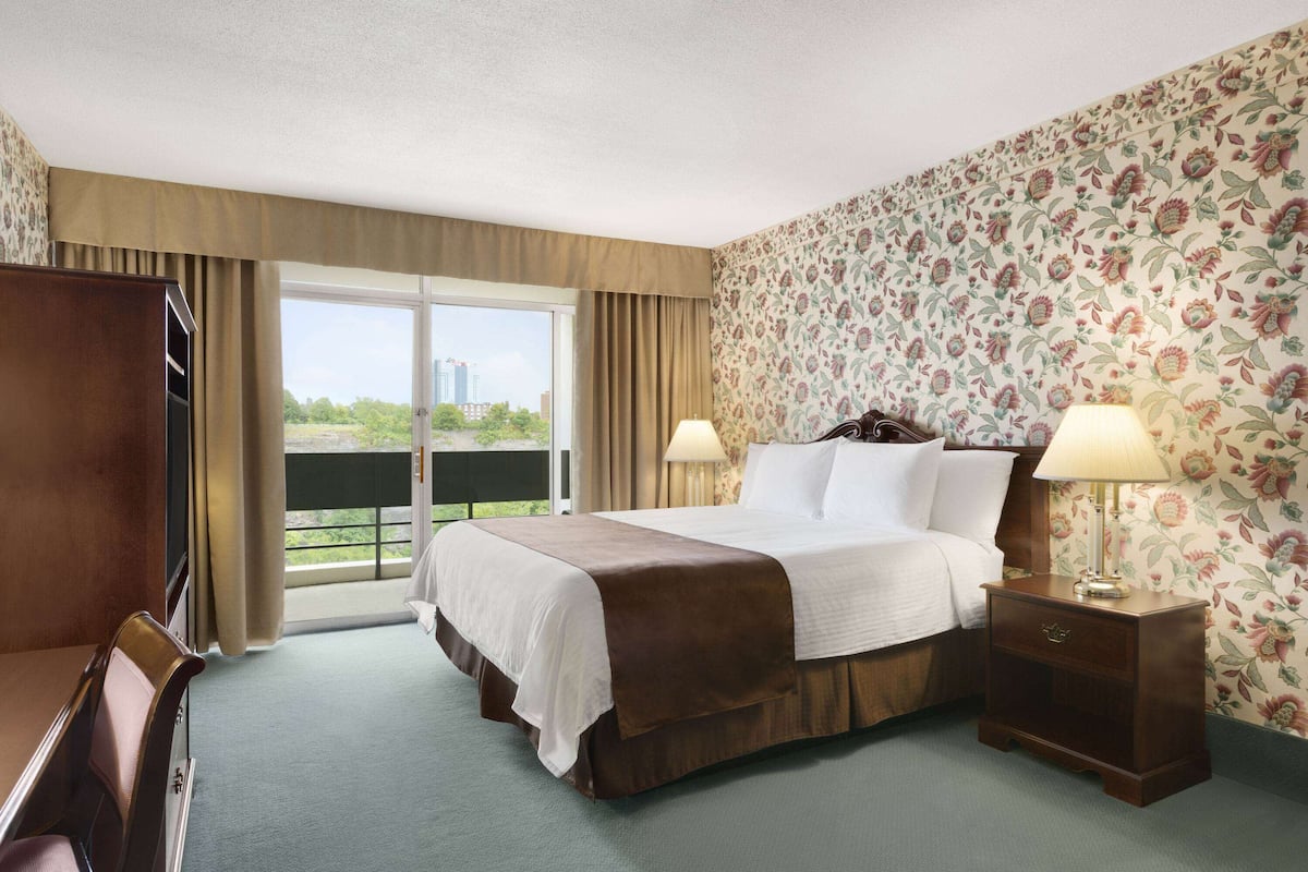 A hotel room with a large bed, two bedside lamps, a wooden nightstand, a desk, and a chair. There is a balcony with a view of buildings and greenery.