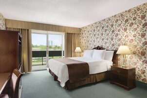 Une chambre d’hôtel avec un grand lit, deux lampes de chevet, une table de chevet en bois, un bureau et une chaise. La chambre dispose d’un balcon avec vue sur les bâtiments et la verdure.