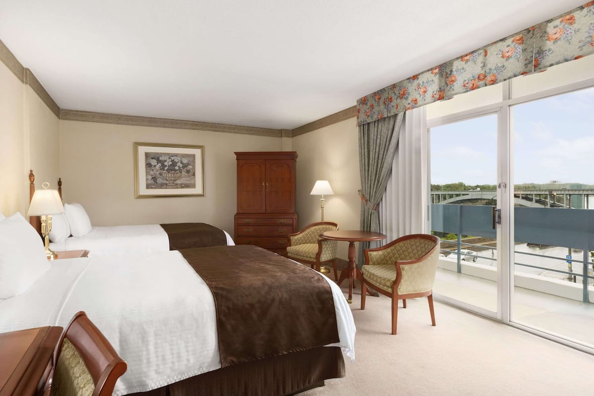 A hotel room with two beds, a desk, a chair, a small table, and a balcony with a view.