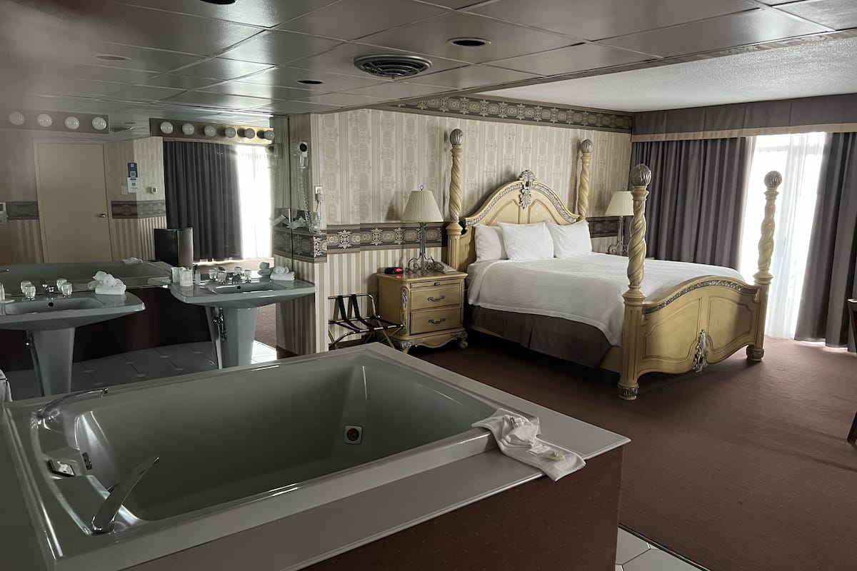 A hotel room with a large bathtub, a four-poster bed, a vanity with a mirror, and a bedside table.