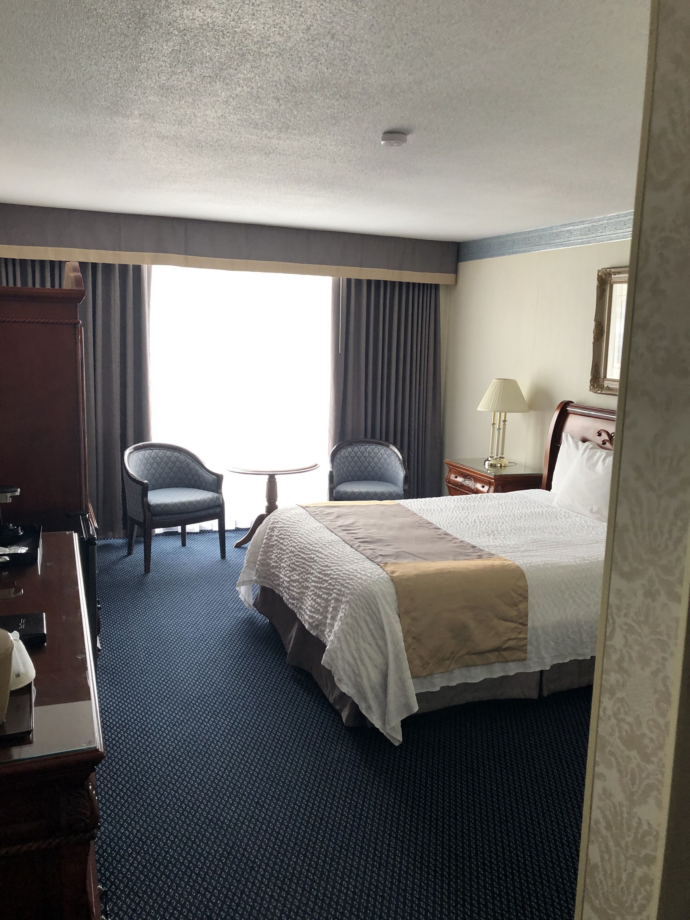 A hotel room with a large bed, two armchairs, a desk with a lamp, and a window with curtains.