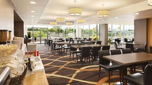 Un moderno comedor de hotel con varias mesas y sillas, una barra de desayuno tipo buffet y grandes ventanales con vistas al exterior.