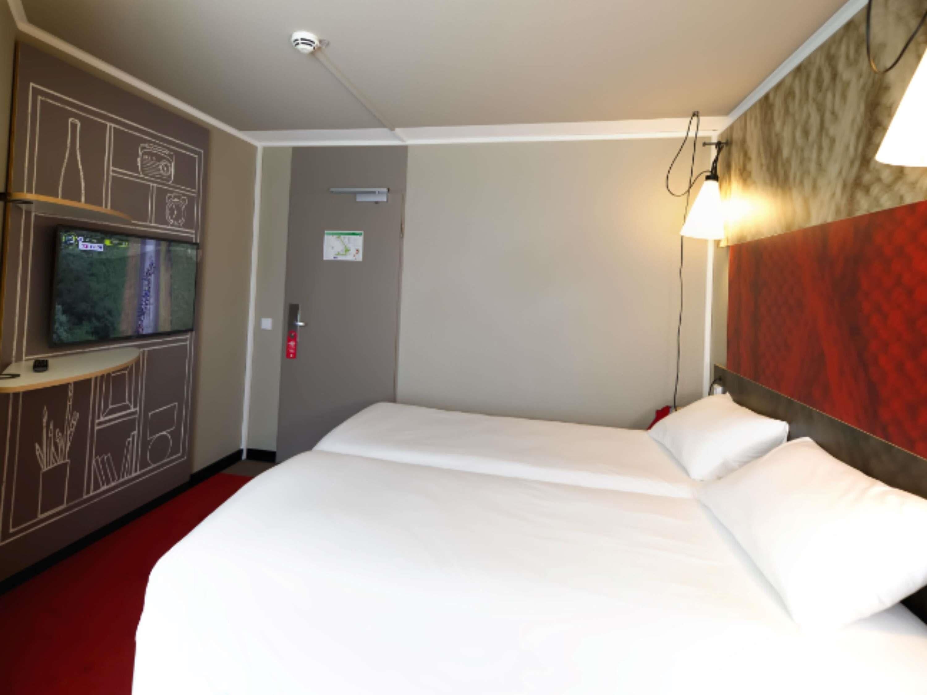 Habitación de hotel con cama, televisor sobre un pedestal, luz instalada en la pared y puerta con detector de incendios.
