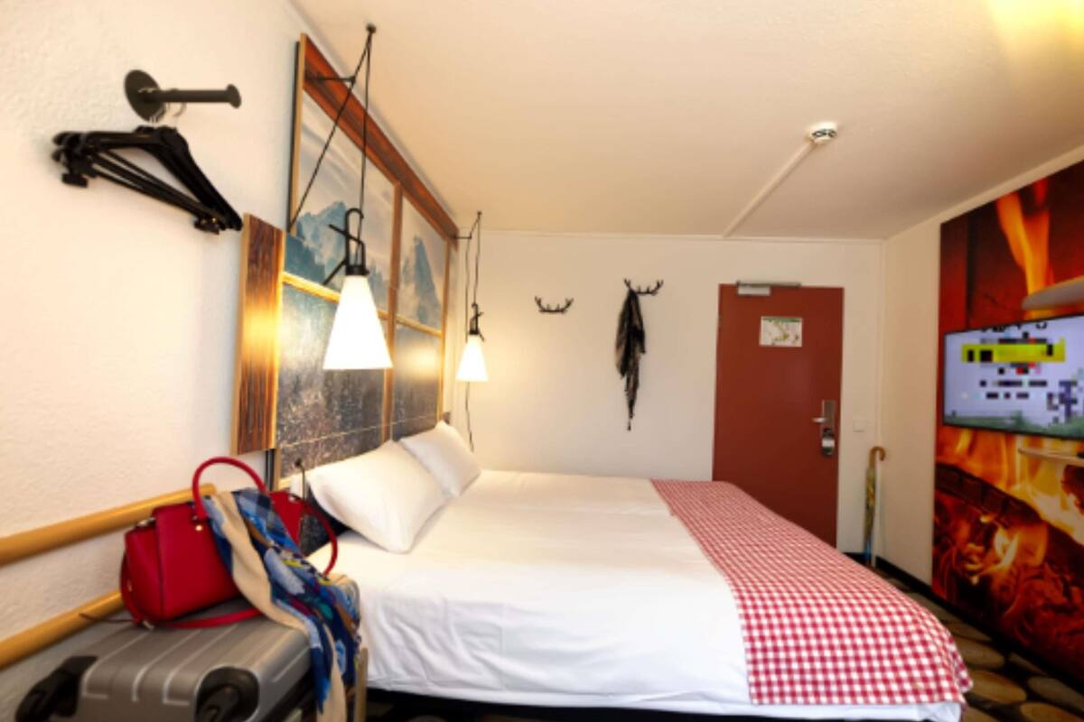 A hotel room with a bed, a red and white checkered blanket, a suitcase, a red handbag, wall-mounted lights, a door, and a wall with a fire-themed artwork.