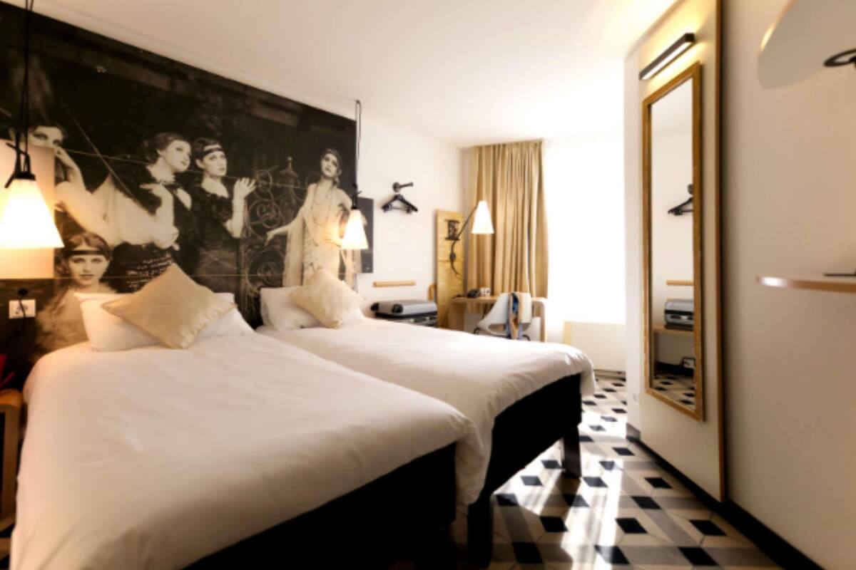 A hotel room with two beds, a large mirror, a painting on the wall, and a check-in counter.