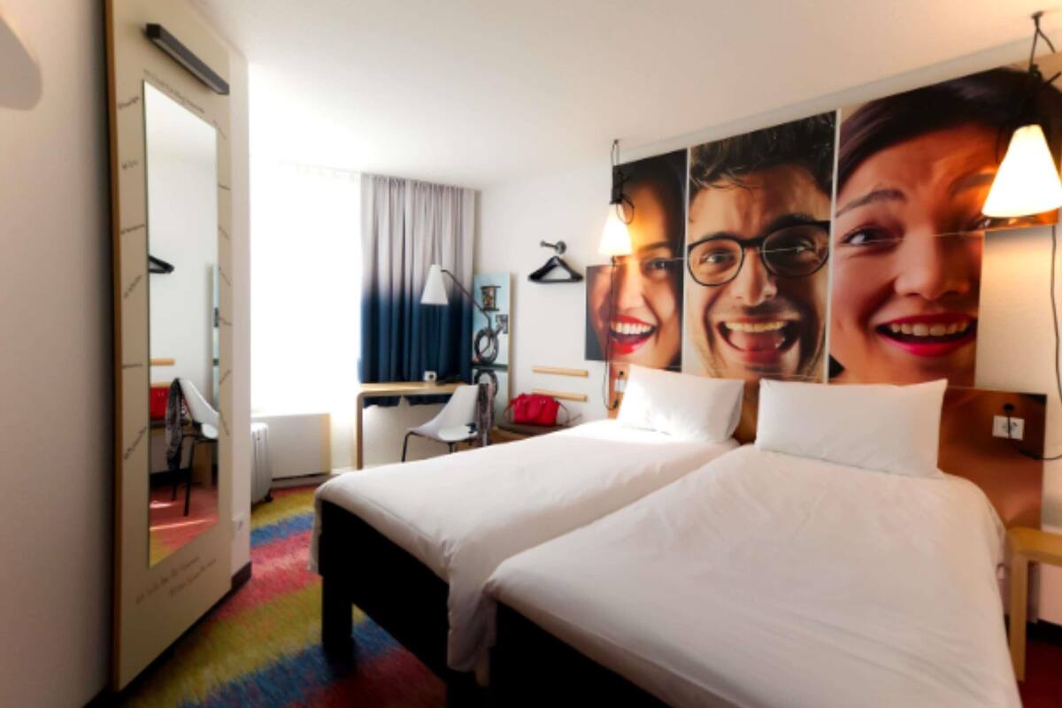 A hotel room with a bed, a desk, a chair, a mirror, and a wall with a large poster.