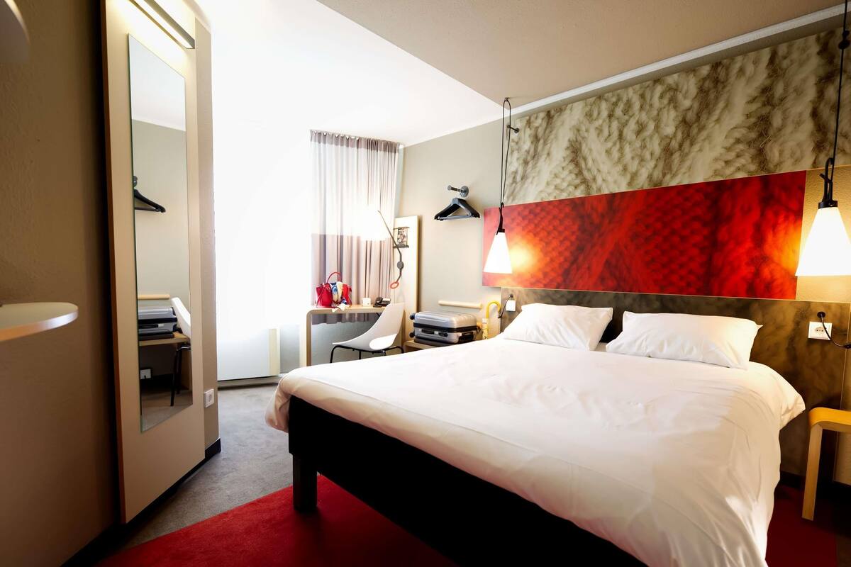 A hotel room with a bed, a desk with a chair, a wardrobe, and a red carpet.