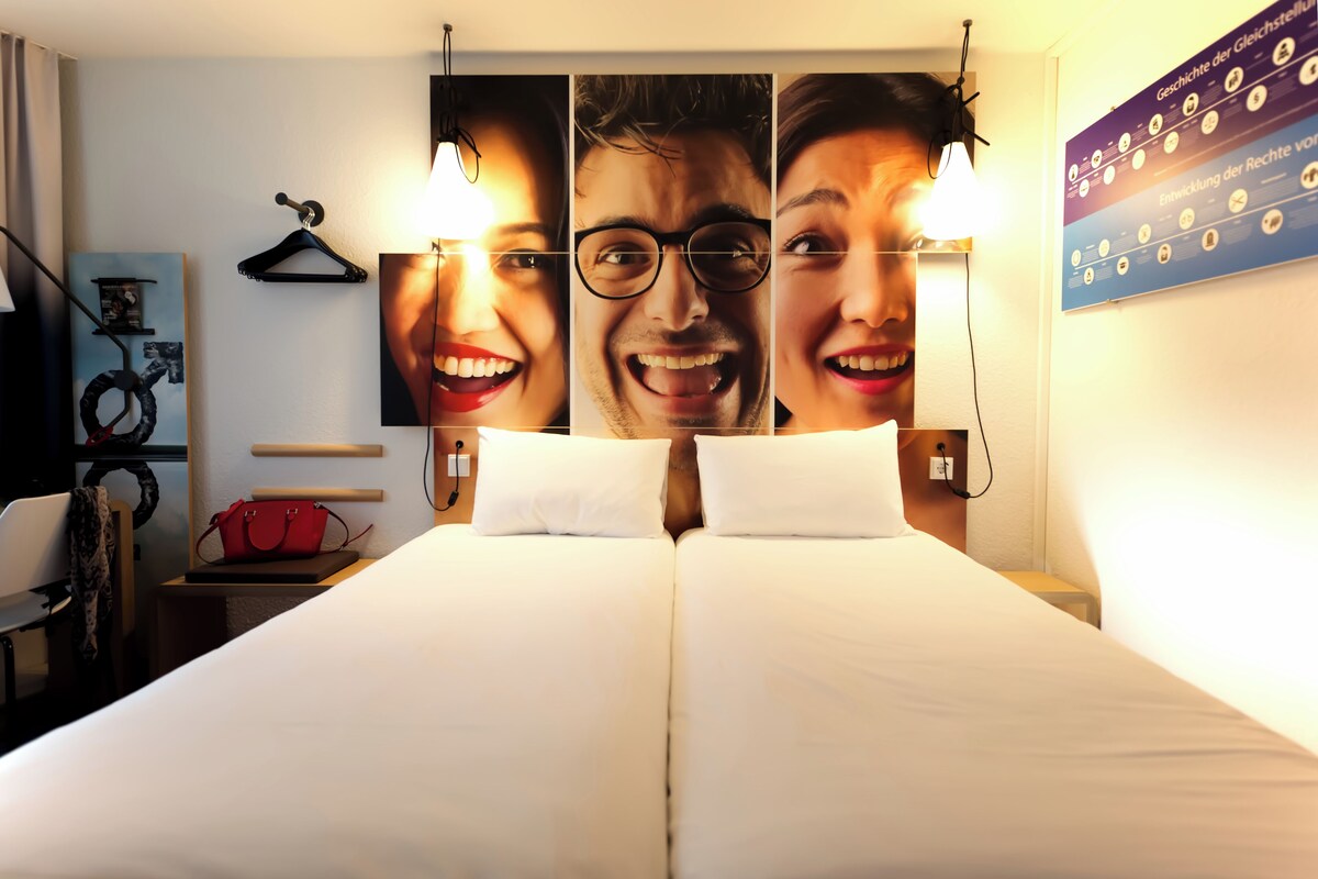 A hotel room with a bed, a red handbag, a wall-mounted light, and a poster.