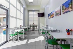 A bright, sunlit dining area with white tables and green chairs, a flat-screen TV on the wall, and framed pictures. - Quality Inn University North I-75 (Gainesville)
