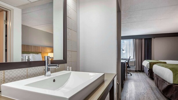 A hotel bathroom with a large sink, a mirror, and a bed visible in the adjacent room.