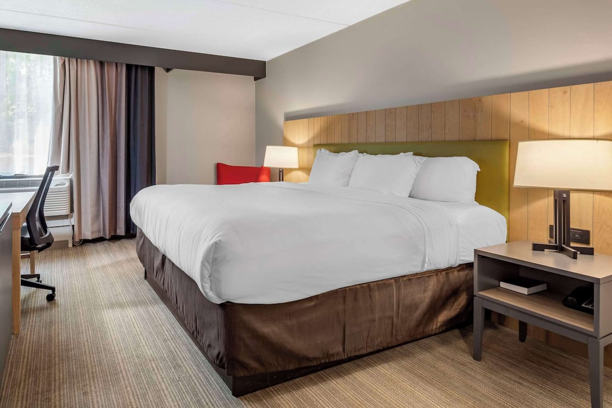 A hotel room with a large bed, a desk with a chair, a lamp, and a window with curtains.