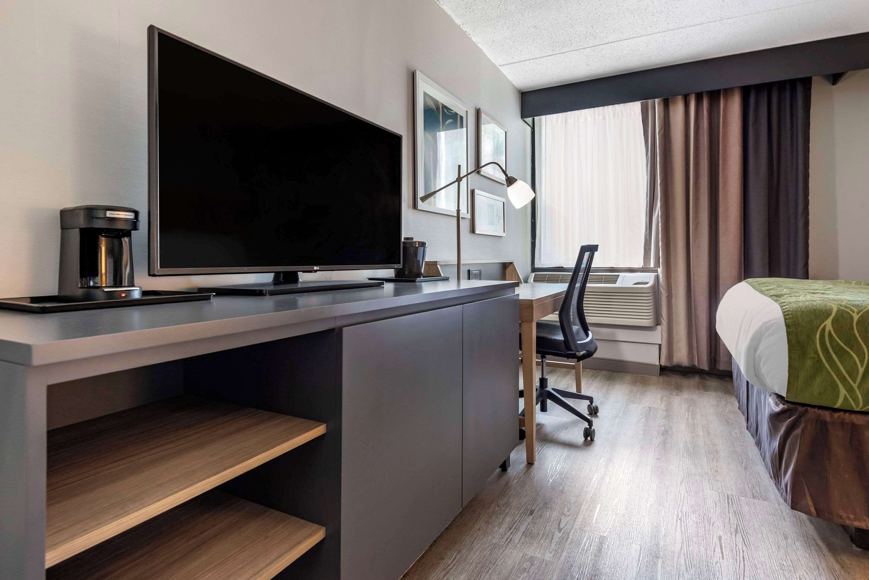 A modern hotel room with a large flat-screen TV, a coffee maker, a desk with a chair, and a bed with a green blanket.