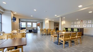 A spacious dining area with multiple wooden tables and chairs, a kitchen area in the background, and framed artwork on the walls.