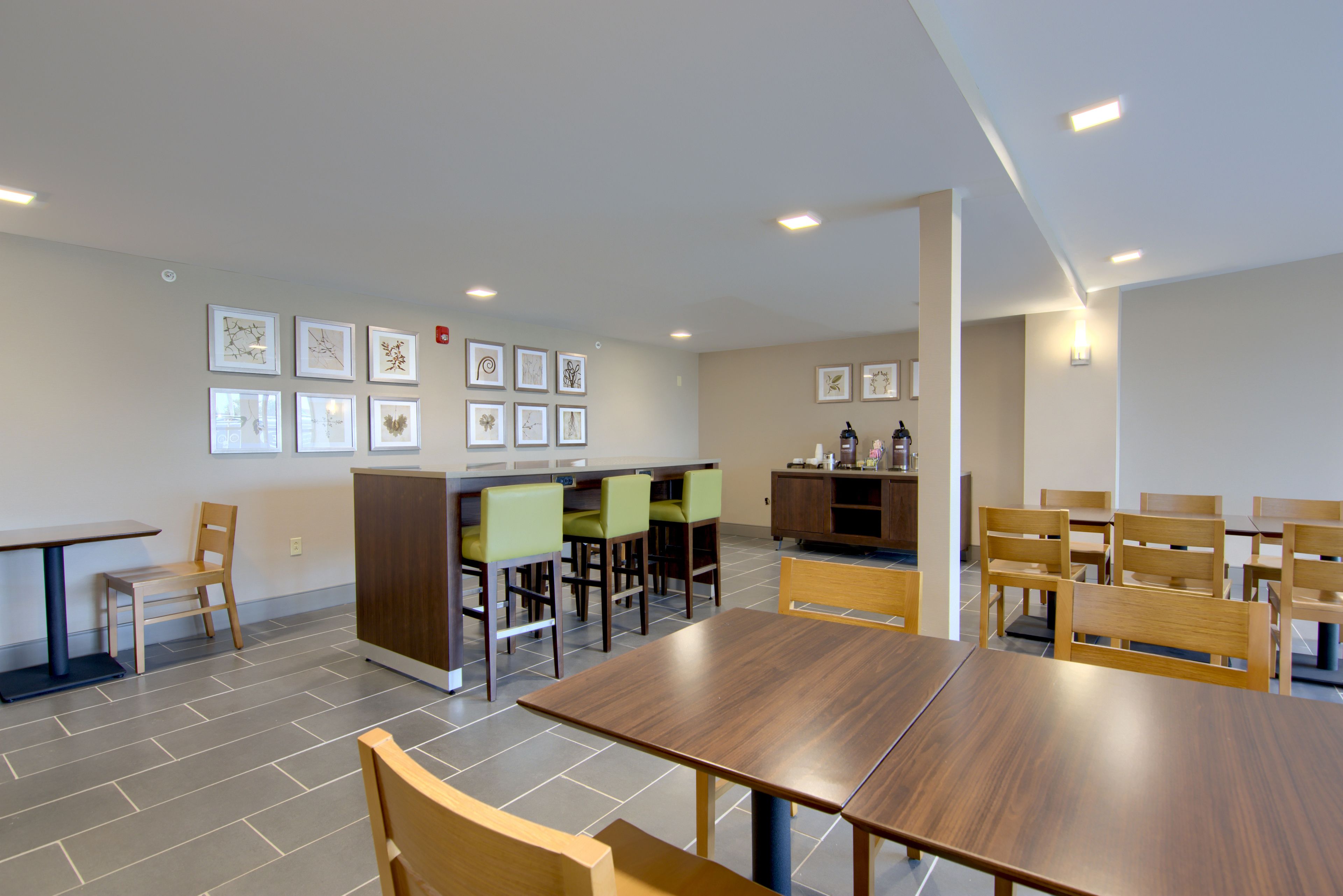 Een moderne eethoek met houten tafels en stoelen, een bar met krukken, en kunstwerken in lijsten aan de muur.
