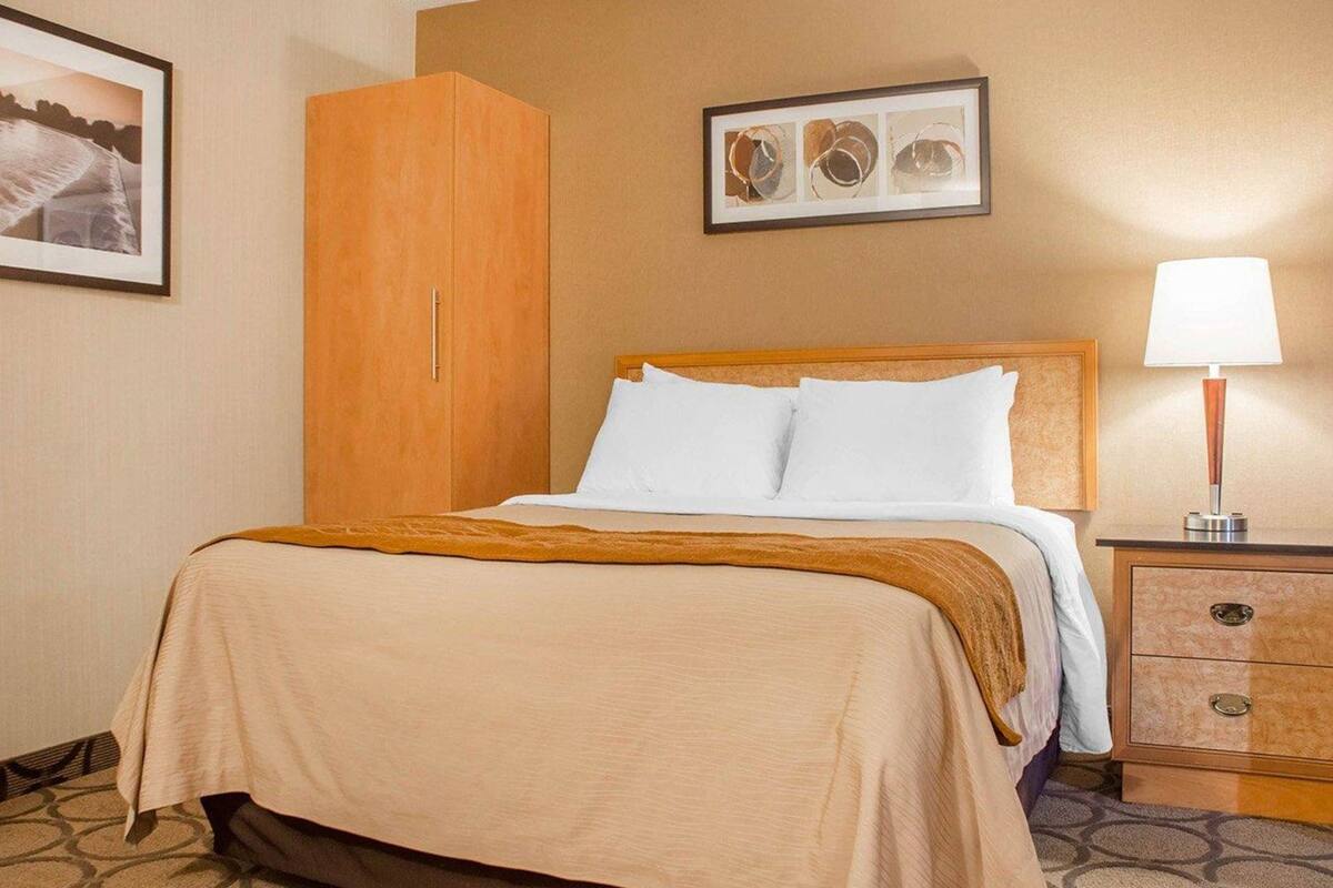 A hotel room with a bed, a wooden wardrobe, a nightstand with a lamp, and a framed picture on the wall.