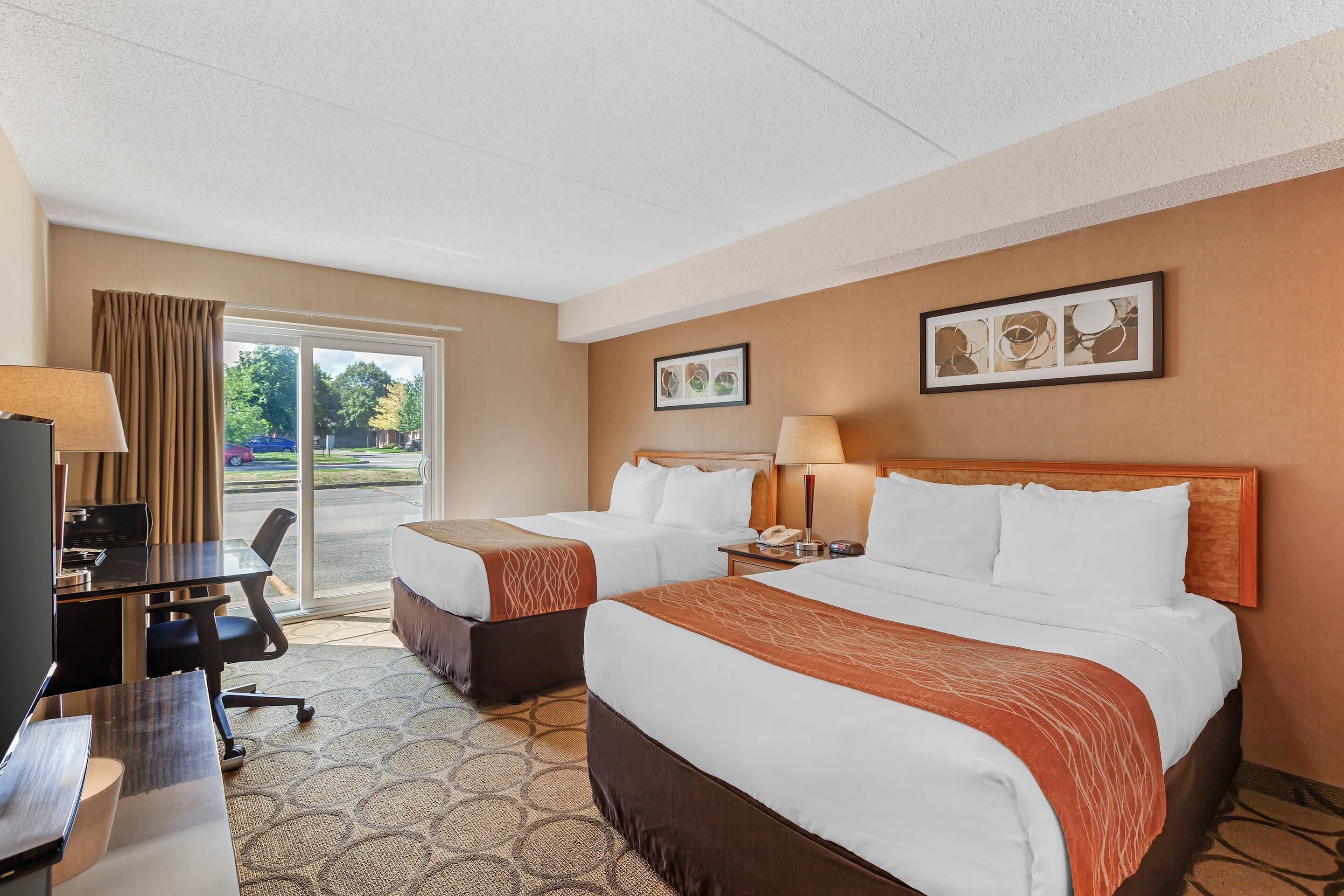 A hotel room with two beds, a desk, and a chair. There is a window with curtains, a door leading outside, and two framed pictures on the wall.