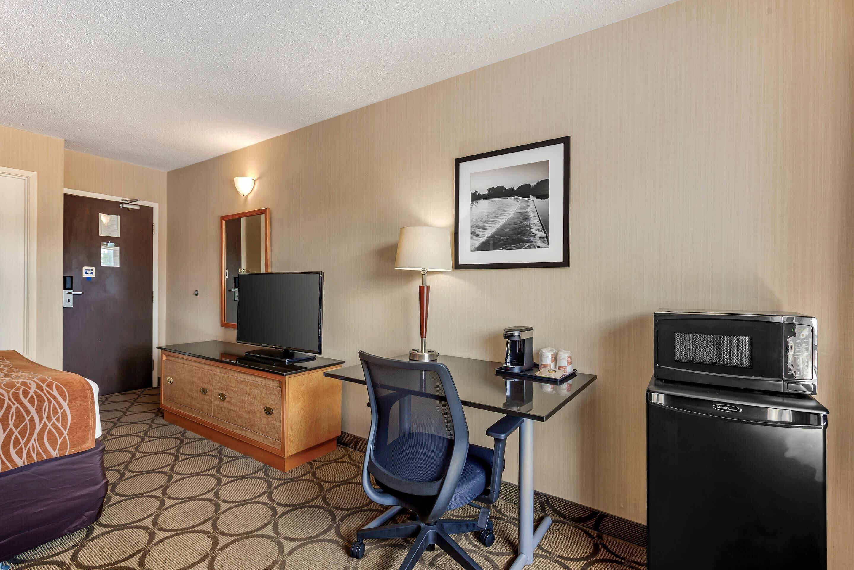 A hotel room with a bed, a desk with a computer, a television, a refrigerator, and a wall-mounted picture.