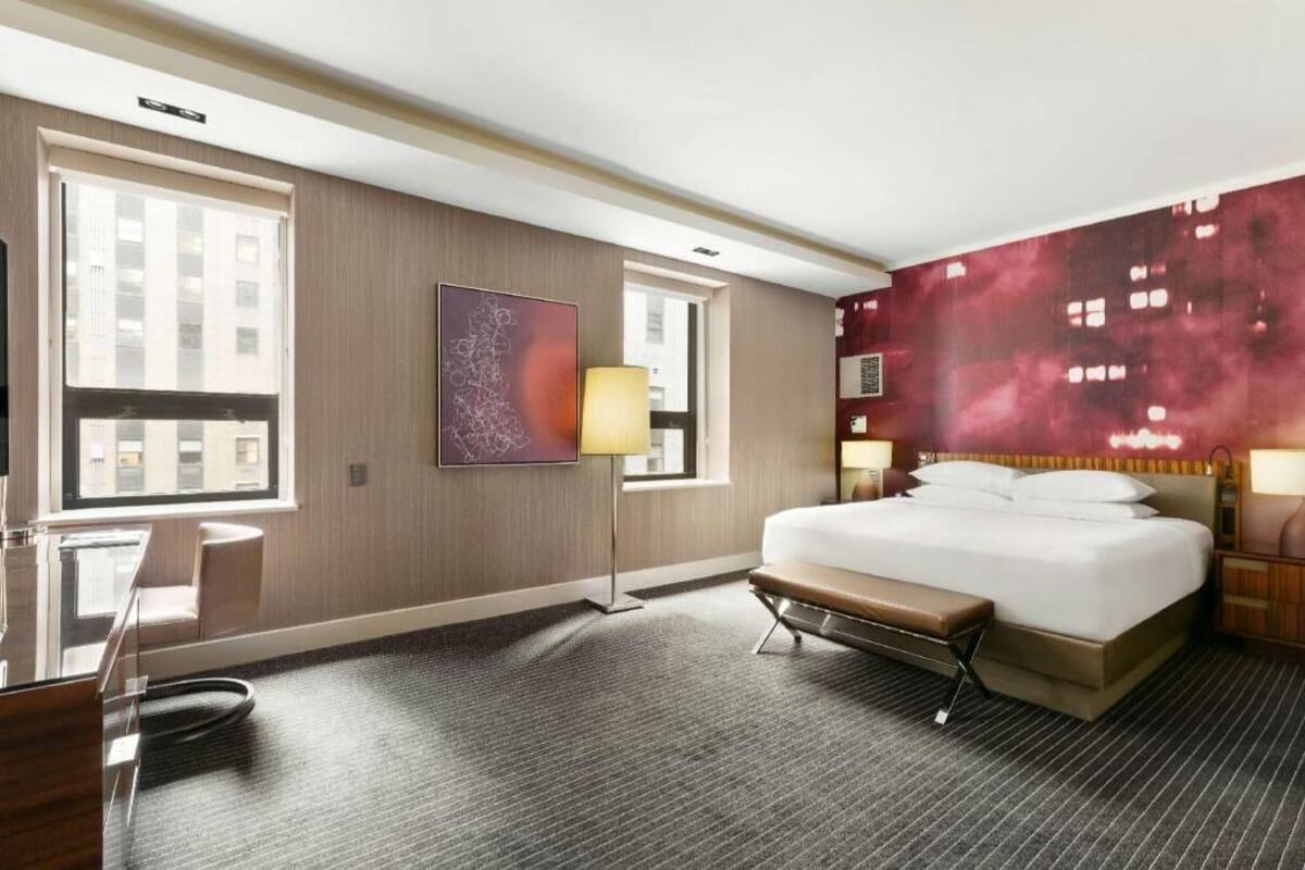 A hotel room with a large bed, a desk, a bench, a window, and a wall with a cityscape mural.