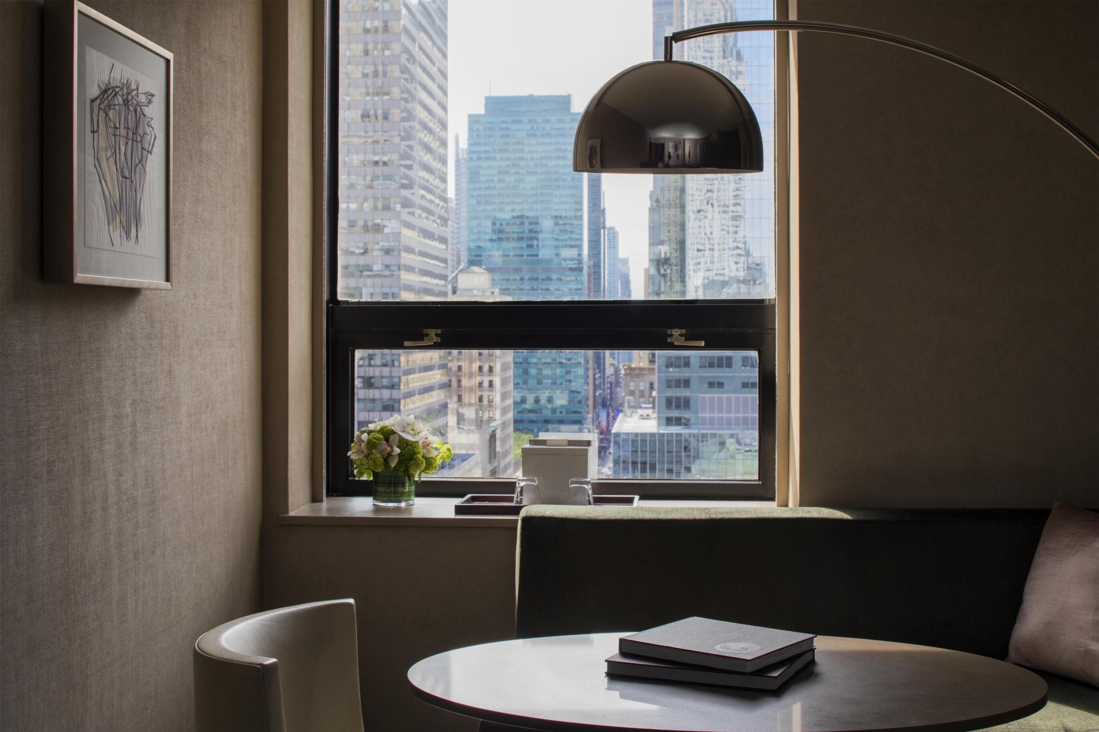 Una habitación moderna con una mesa redonda, una silla, una lámpara de pie, una ventana con vista a edificios de la ciudad y una obra de arte enmarcada en la pared.