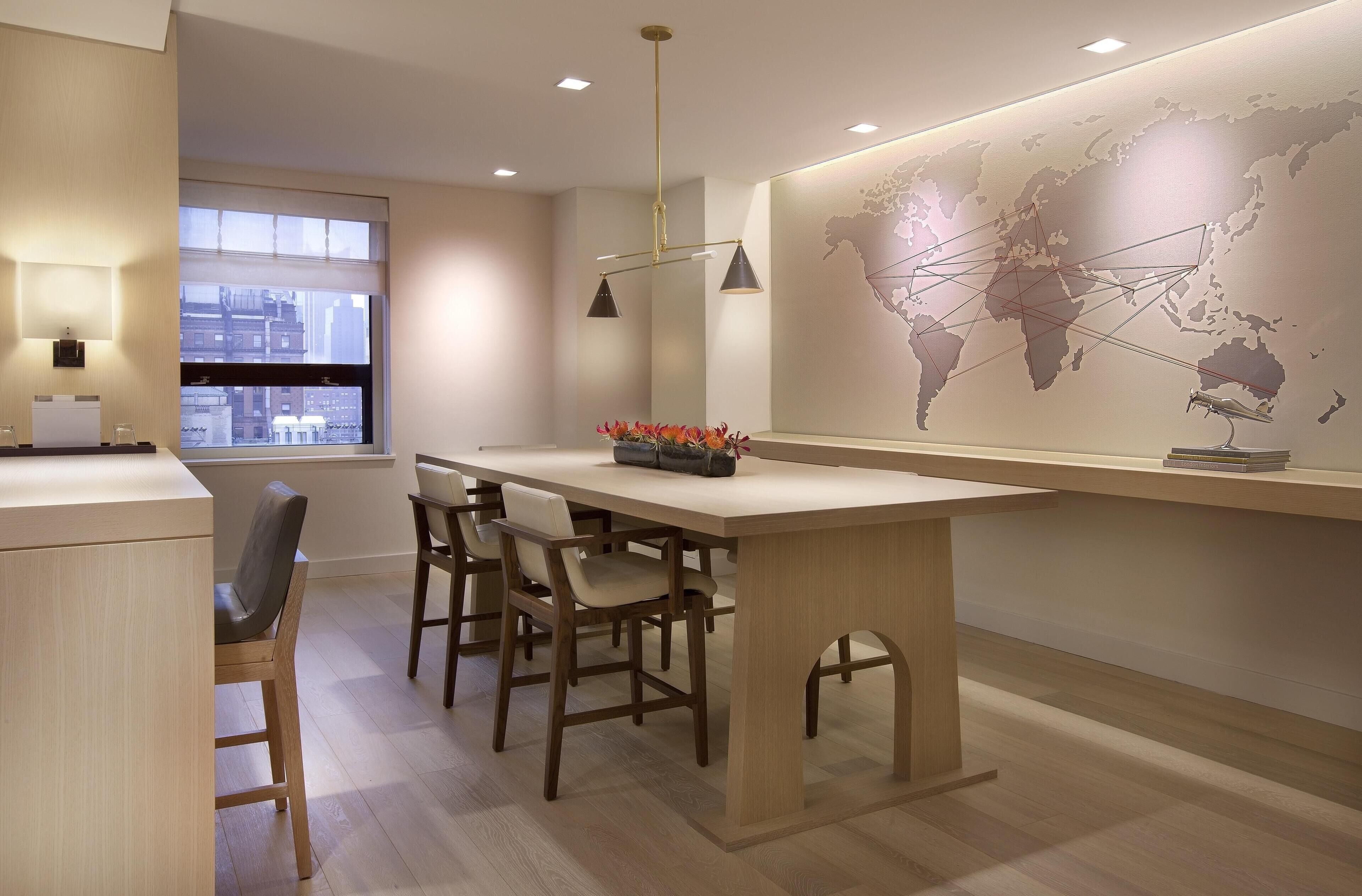 A modern kitchen with a wooden table, bar stools, a world map on the wall, and a window with a city view.