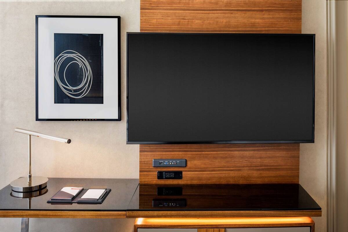A modern hotel room with a large flat-screen TV mounted on a wooden panel, a framed abstract artwork, a sleek desk with a lamp, and a control panel below the TV.