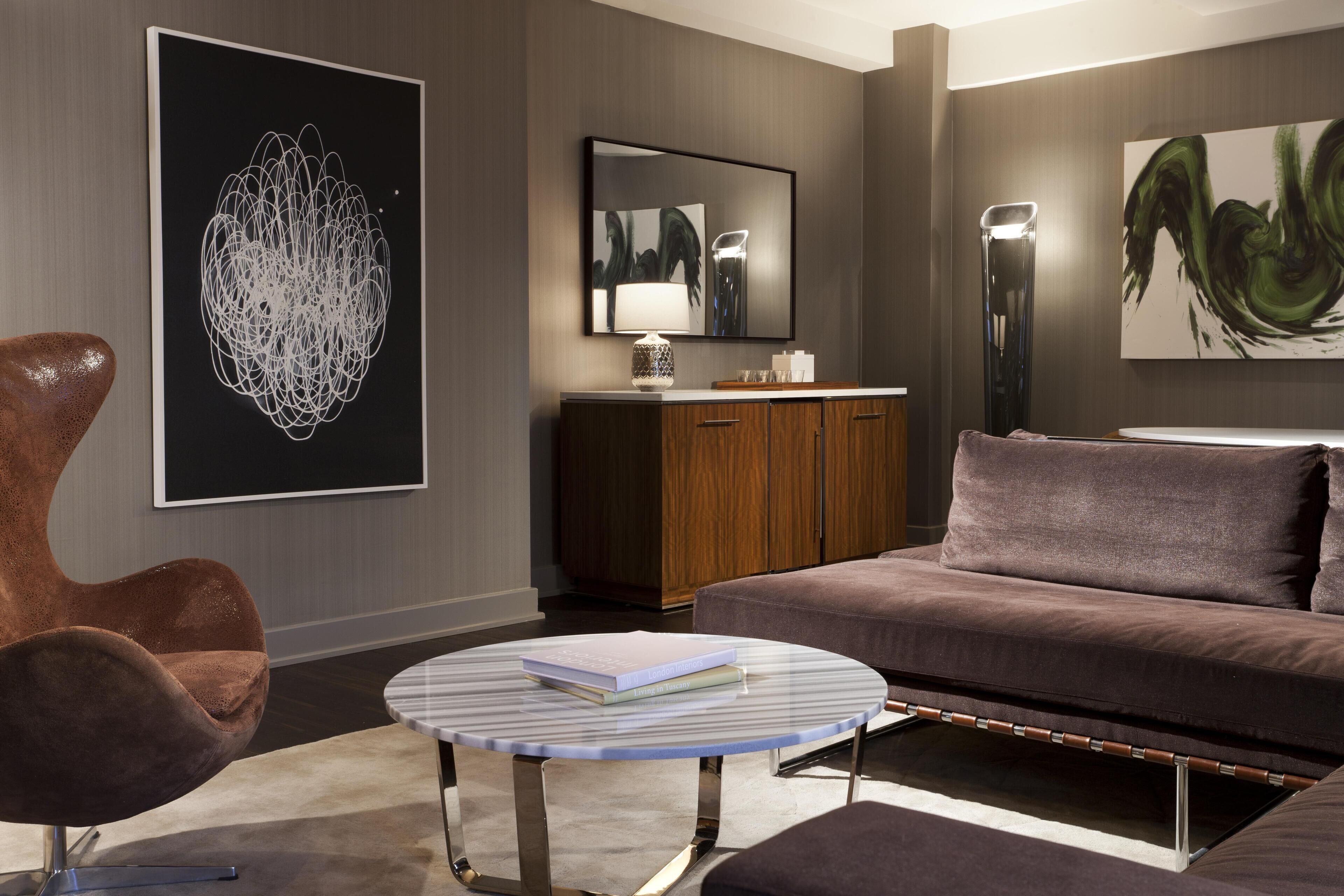 A modern living room with a sofa, a round coffee table, and a chair. There are artworks on the walls and a cabinet with a mirror.