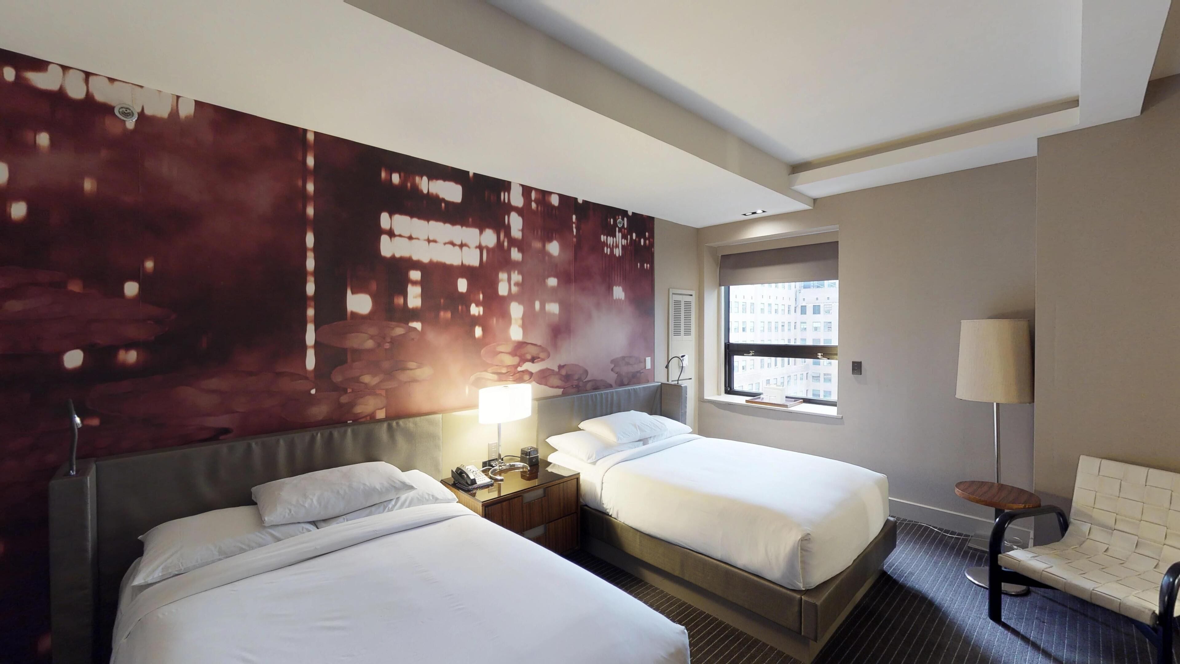 Habitación de hotel con dos camas, un mural grande en la pared, una ventana con vistas a edificios, una mesita de noche con lámpara y una silla.