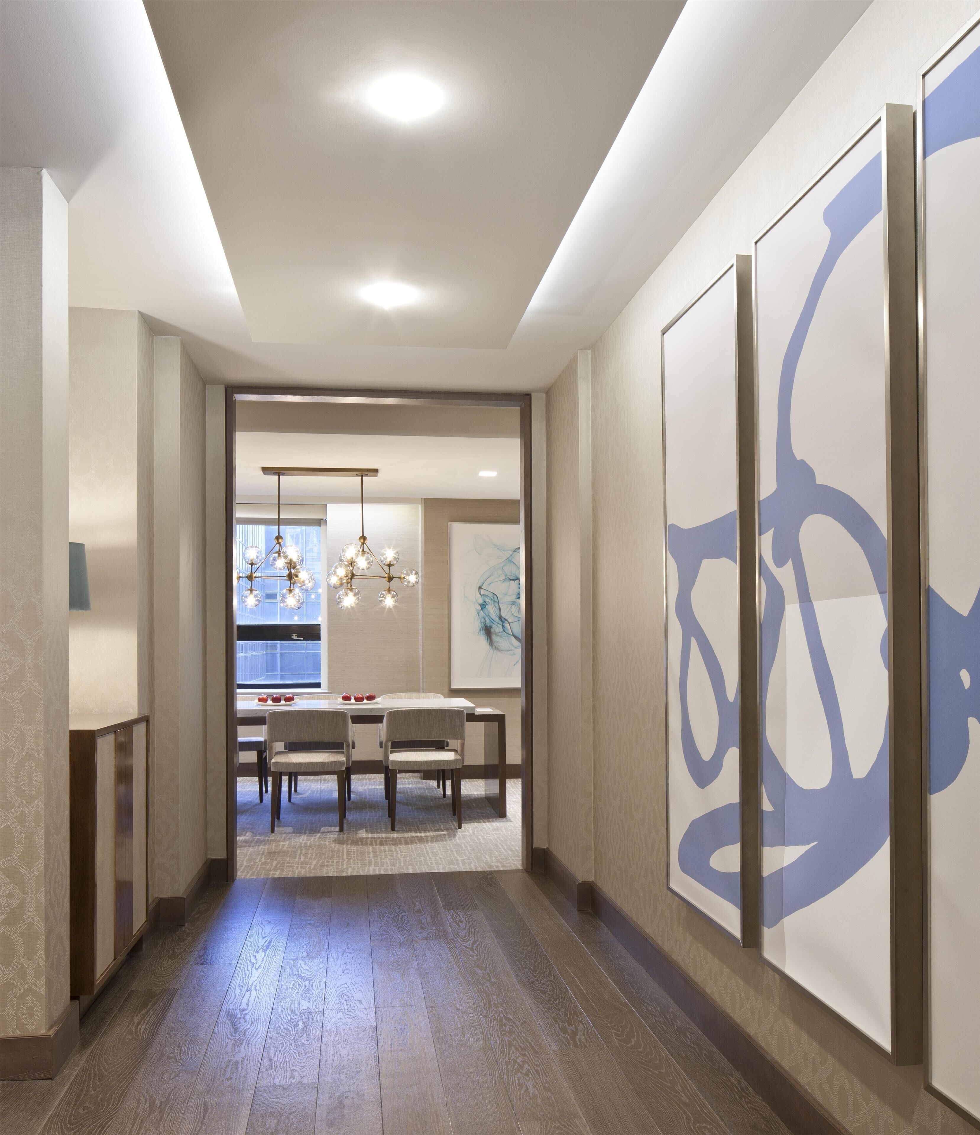 A modern interior with a hallway leading to a dining area, featuring a wooden floor, recessed ceiling lighting, and abstract wall art.