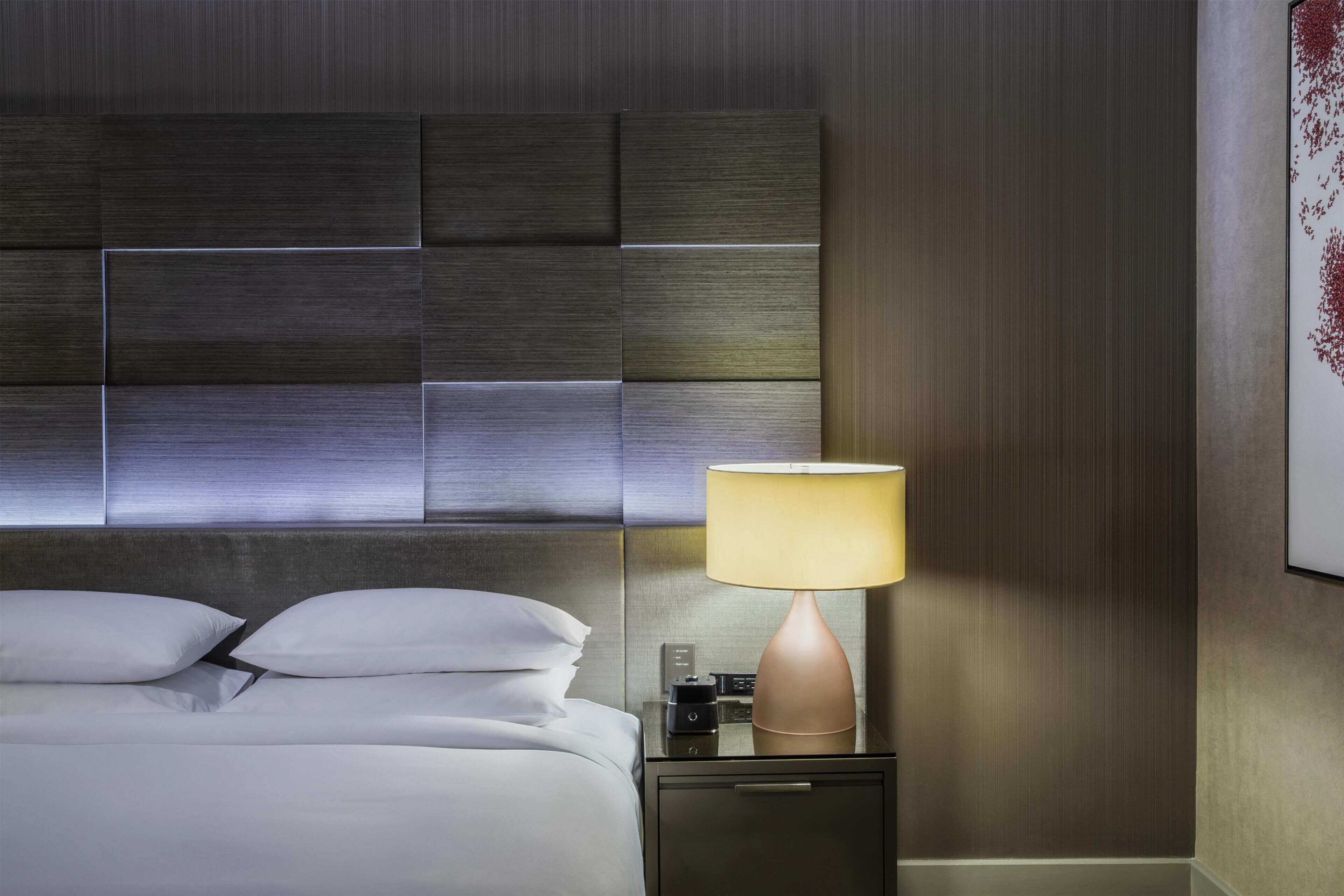 A hotel room with a modern bed, a bedside table with a lamp, and a wall-mounted artwork.