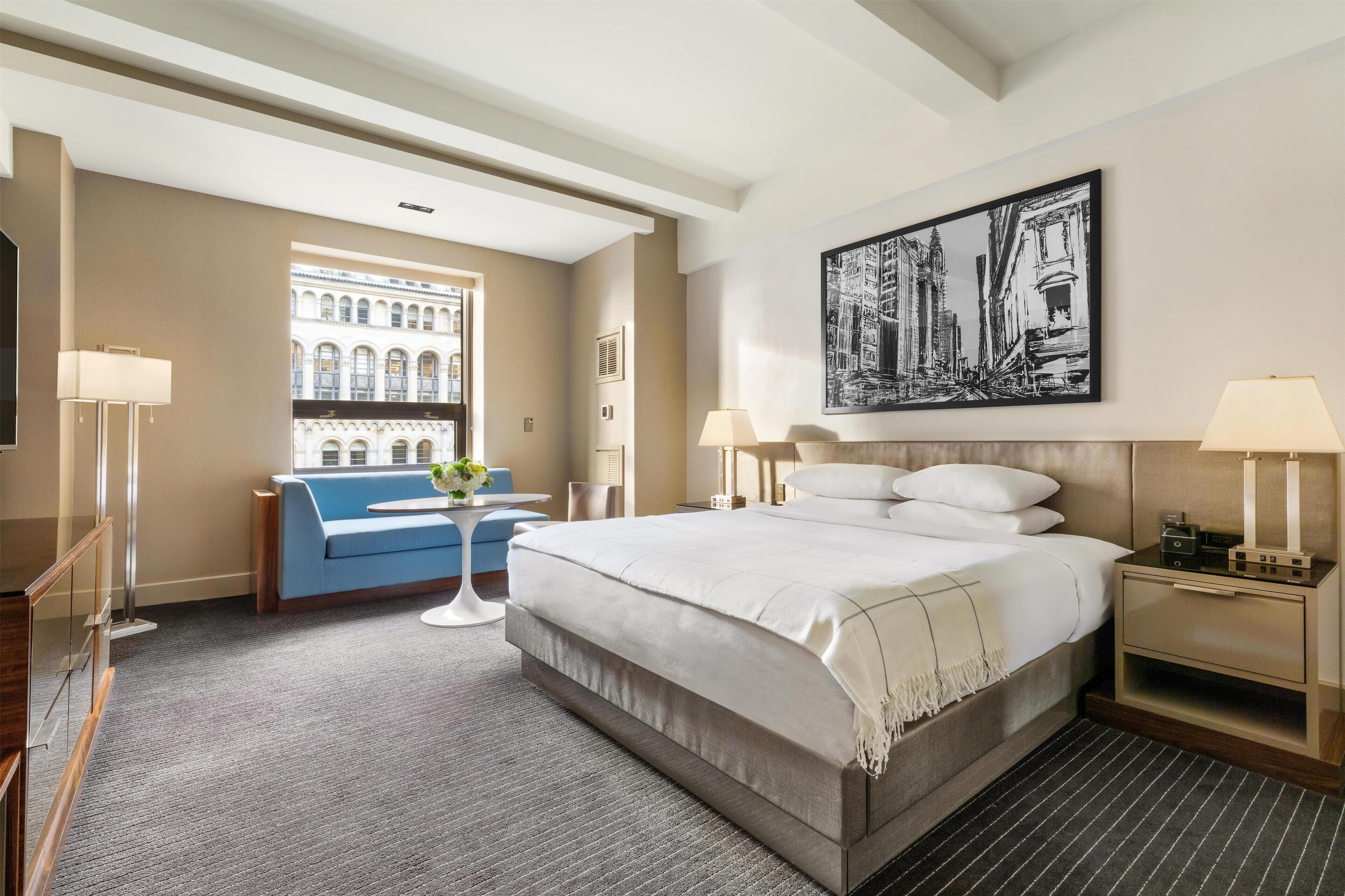 A hotel room with a large bed, a blue sofa, a small table with a vase, and a painting on the wall.