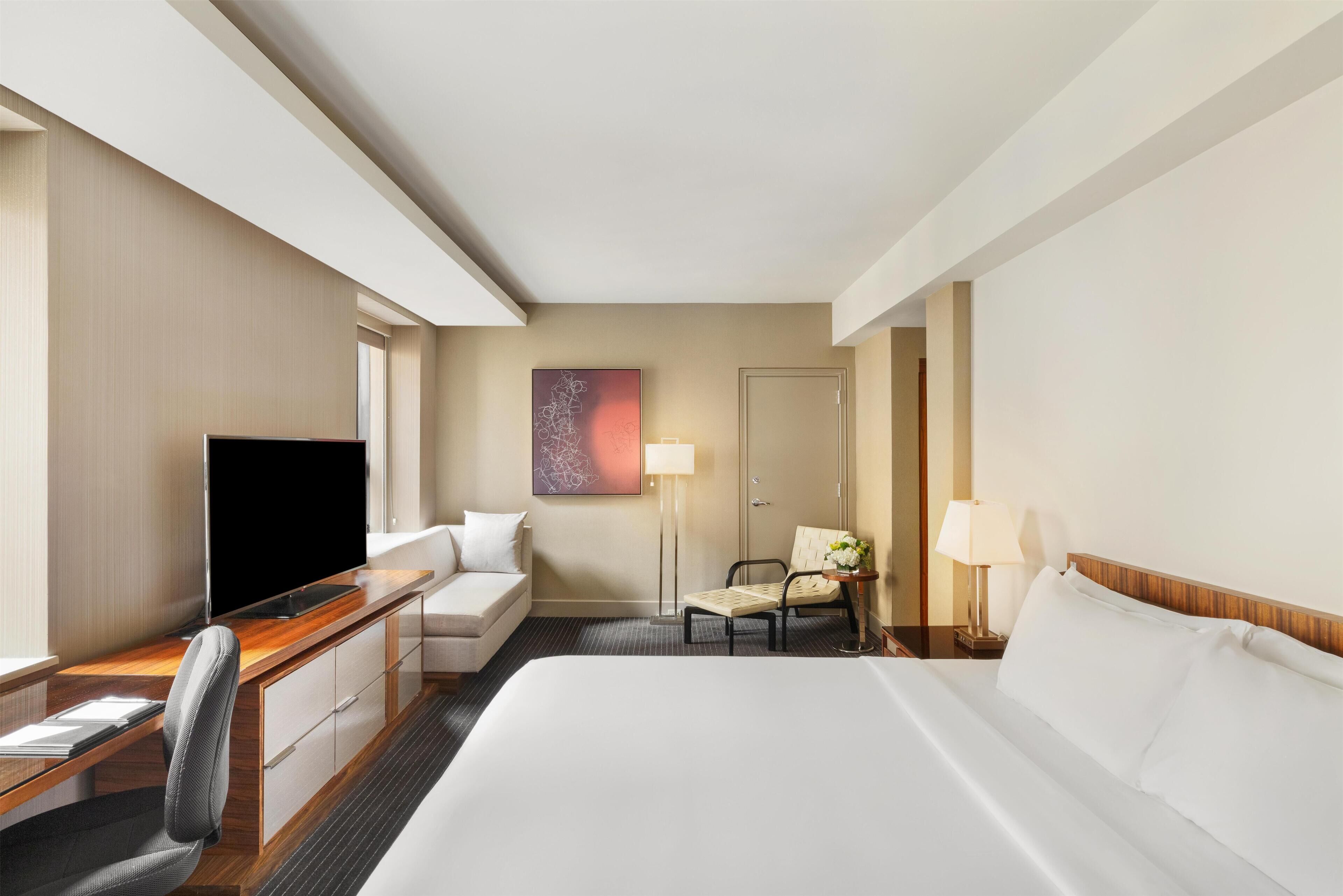 A hotel room with a bed, a desk with a flat-screen TV, a sofa, a chair, a lamp, and a decorative wall piece.