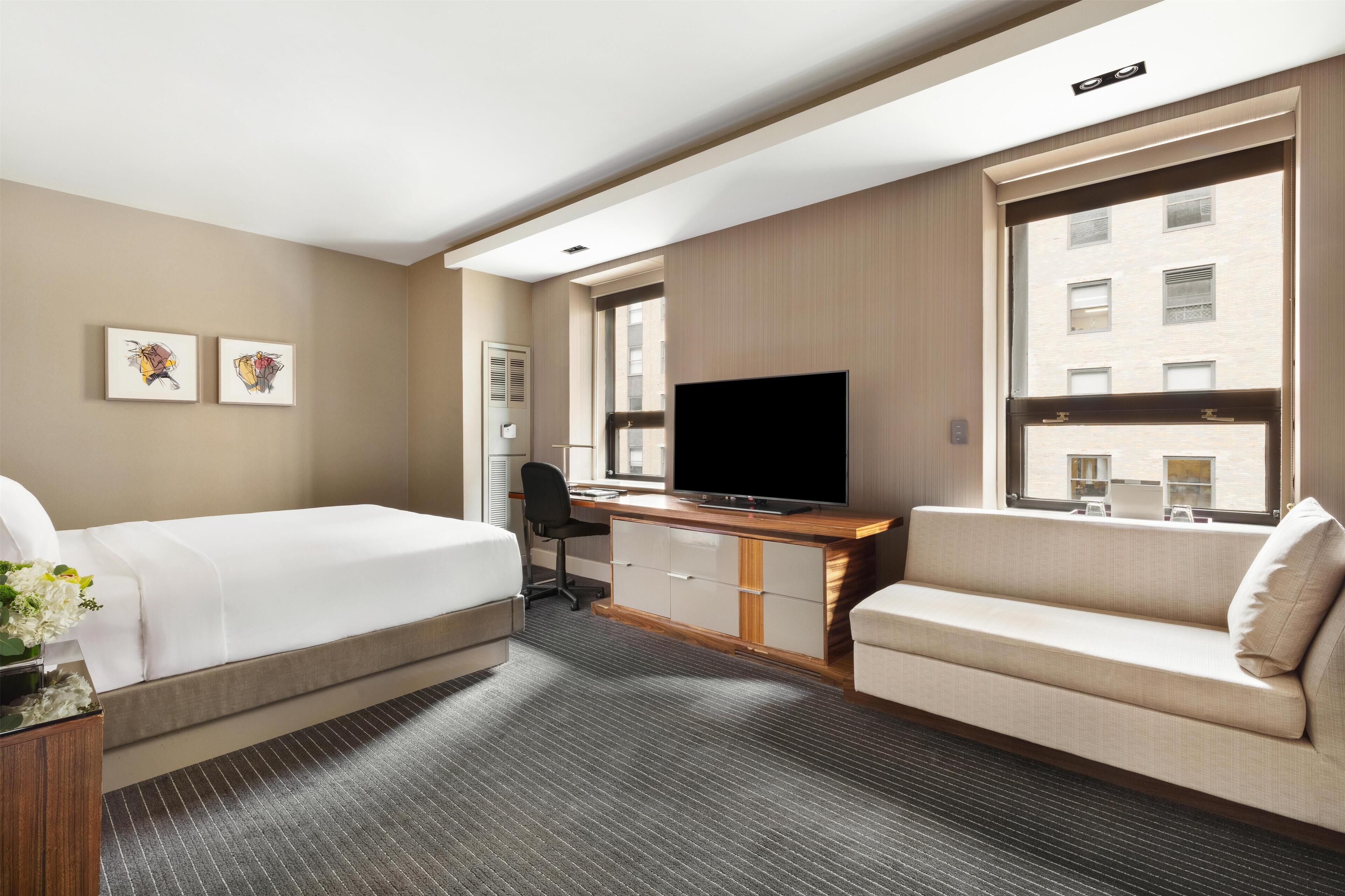 A hotel room with a bed, a desk with a TV, a sofa, and a window with a view of buildings.