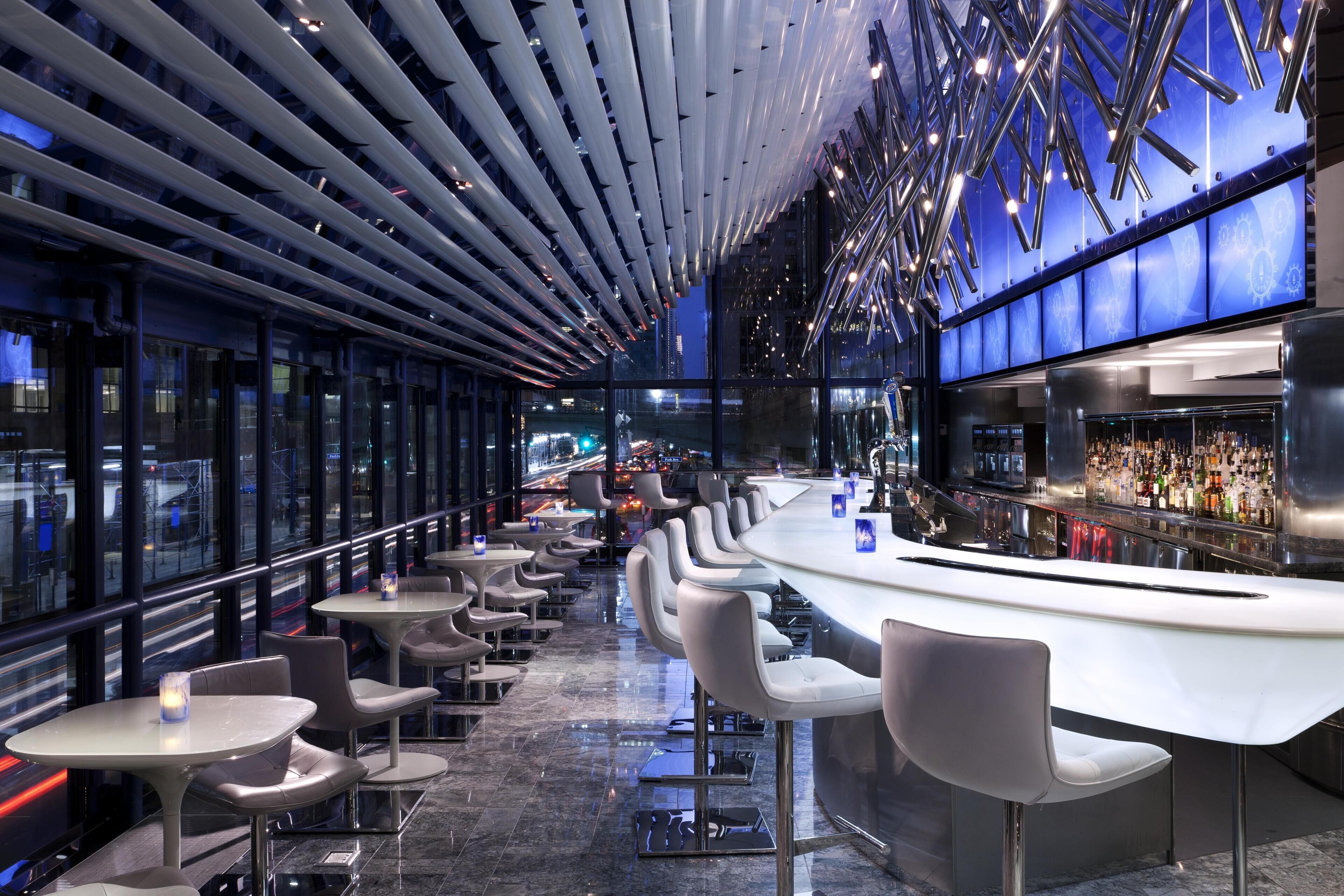 A modern bar with a curved counter, multiple seating areas, and a view of the cityscape through large windows.