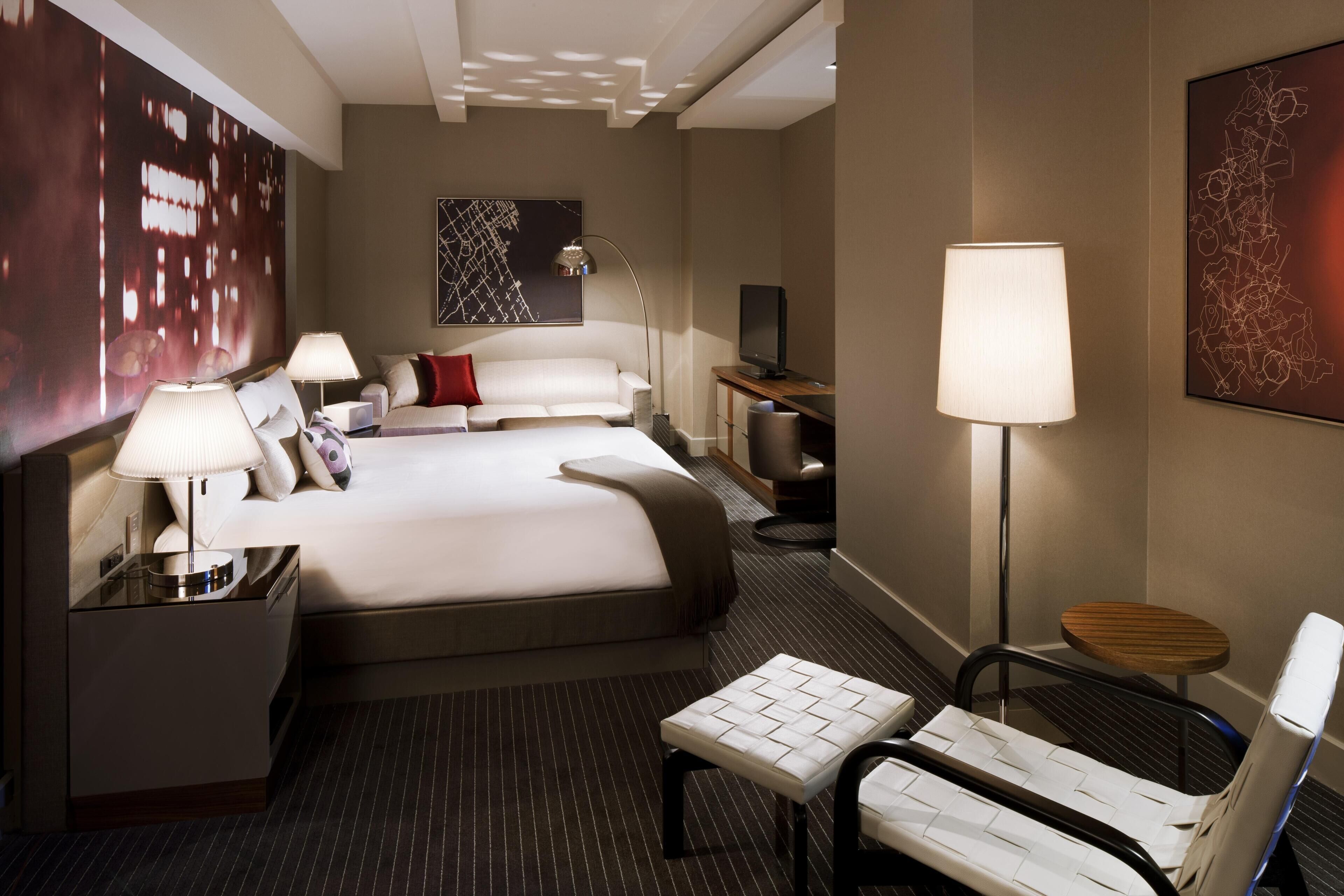Quarto de hotel com uma cama grande, mesas de cabeceira, escrivaninha, cadeira, televisão e obras de arte nas paredes.