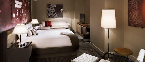 A hotel room with a large bed, bedside tables, a desk, a chair, a TV, and artwork on the walls.