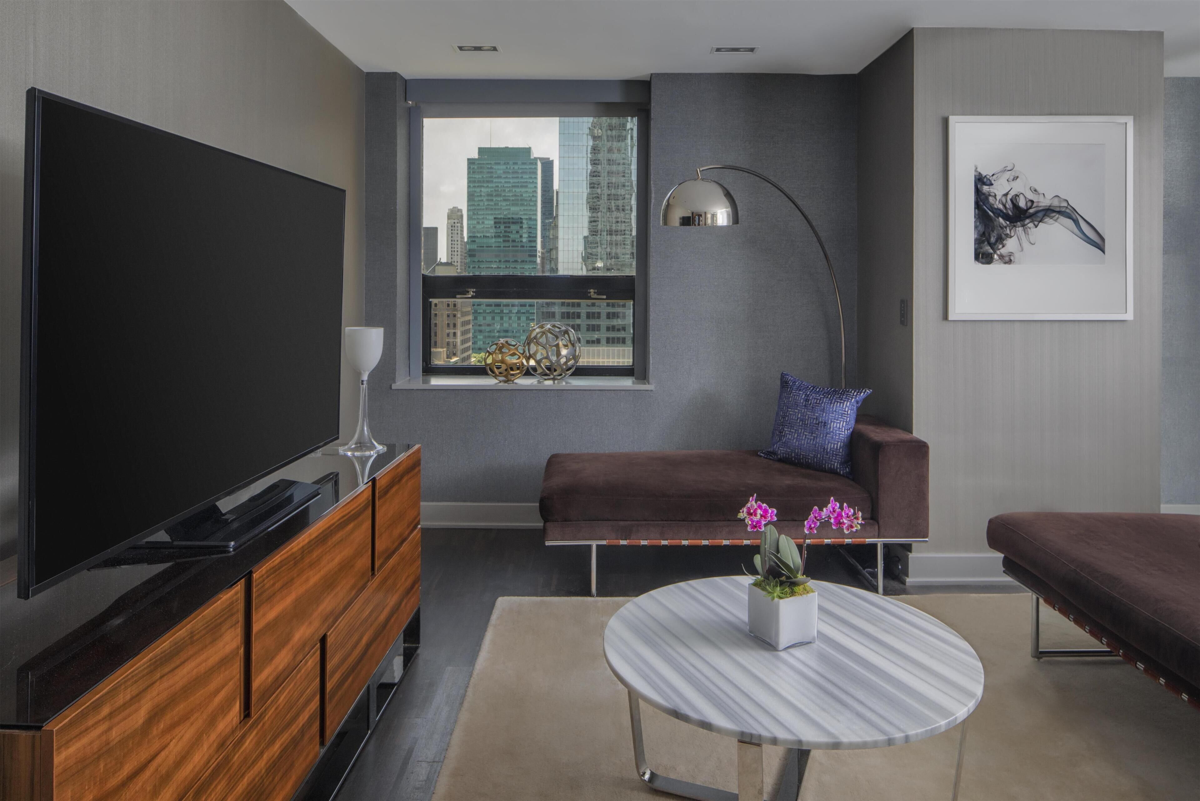 A modern living room with a large flat-screen TV, a sofa, a coffee table with flowers, and a city view through a window.