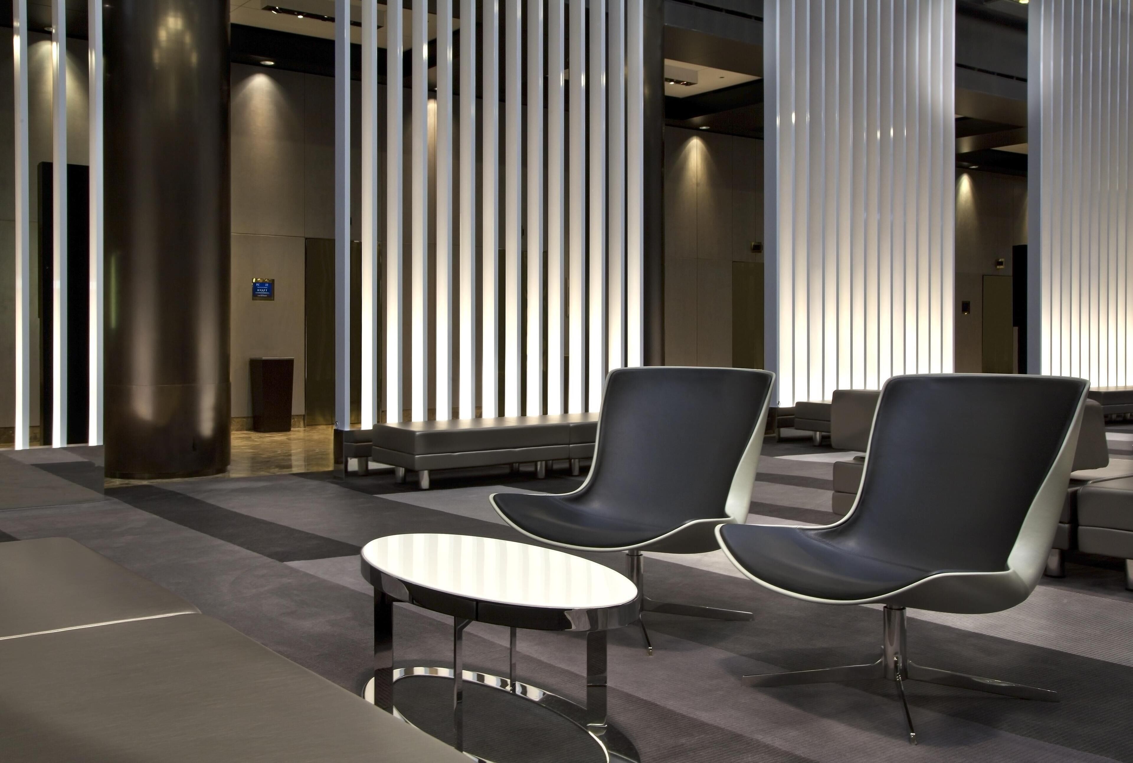 Um lobby moderno com duas cadeiras pretas, uma pequena mesa redonda e painéis de luz verticais.